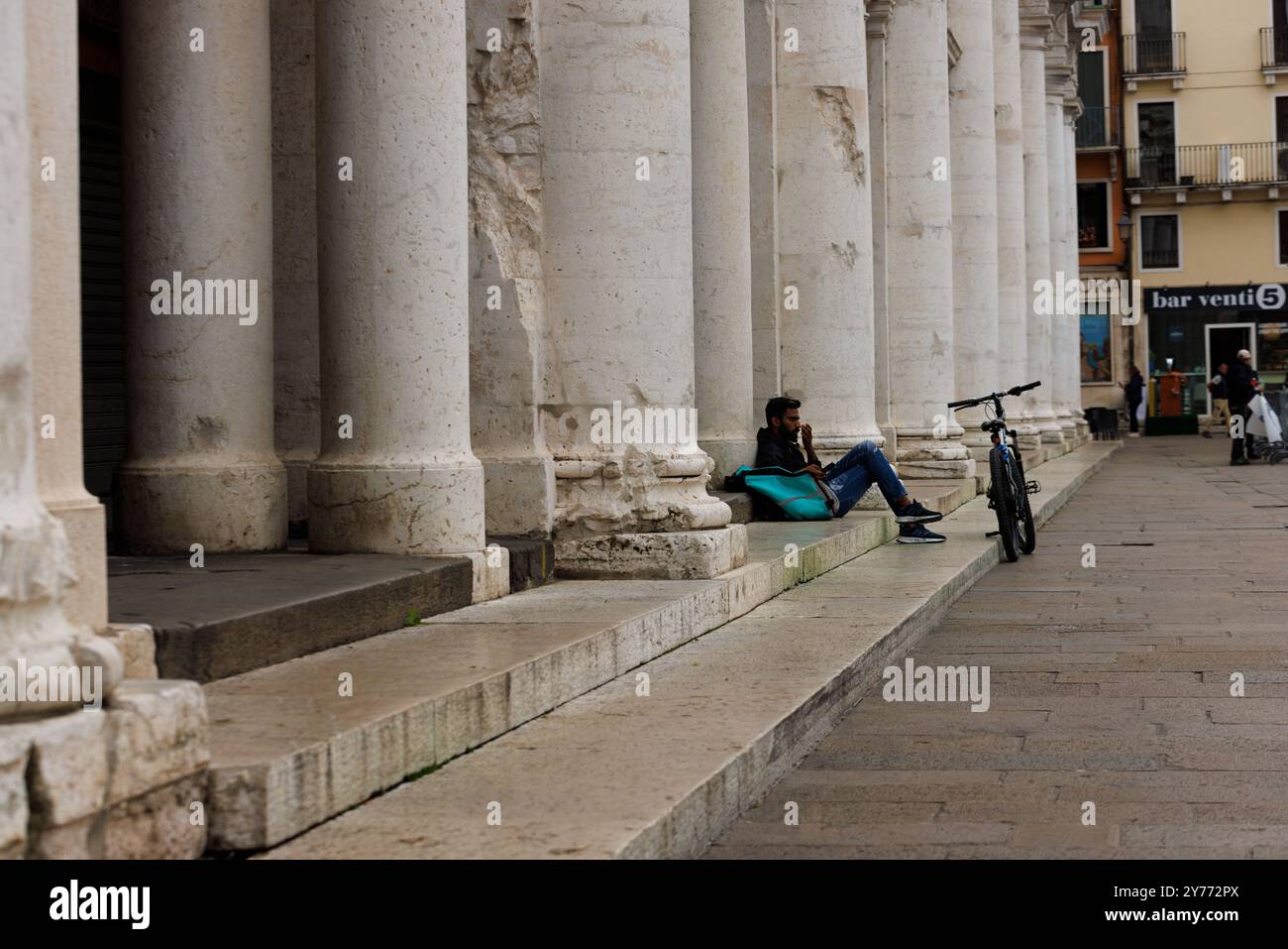 Vicenza, Italien - 13. September 2024: Ein Arbeiter aus Deliveroo macht eine Pause, sitzt auf der Treppe und lehnt sich an seine Arbeitstasche, während sein Fahrrad ruht Stockfoto