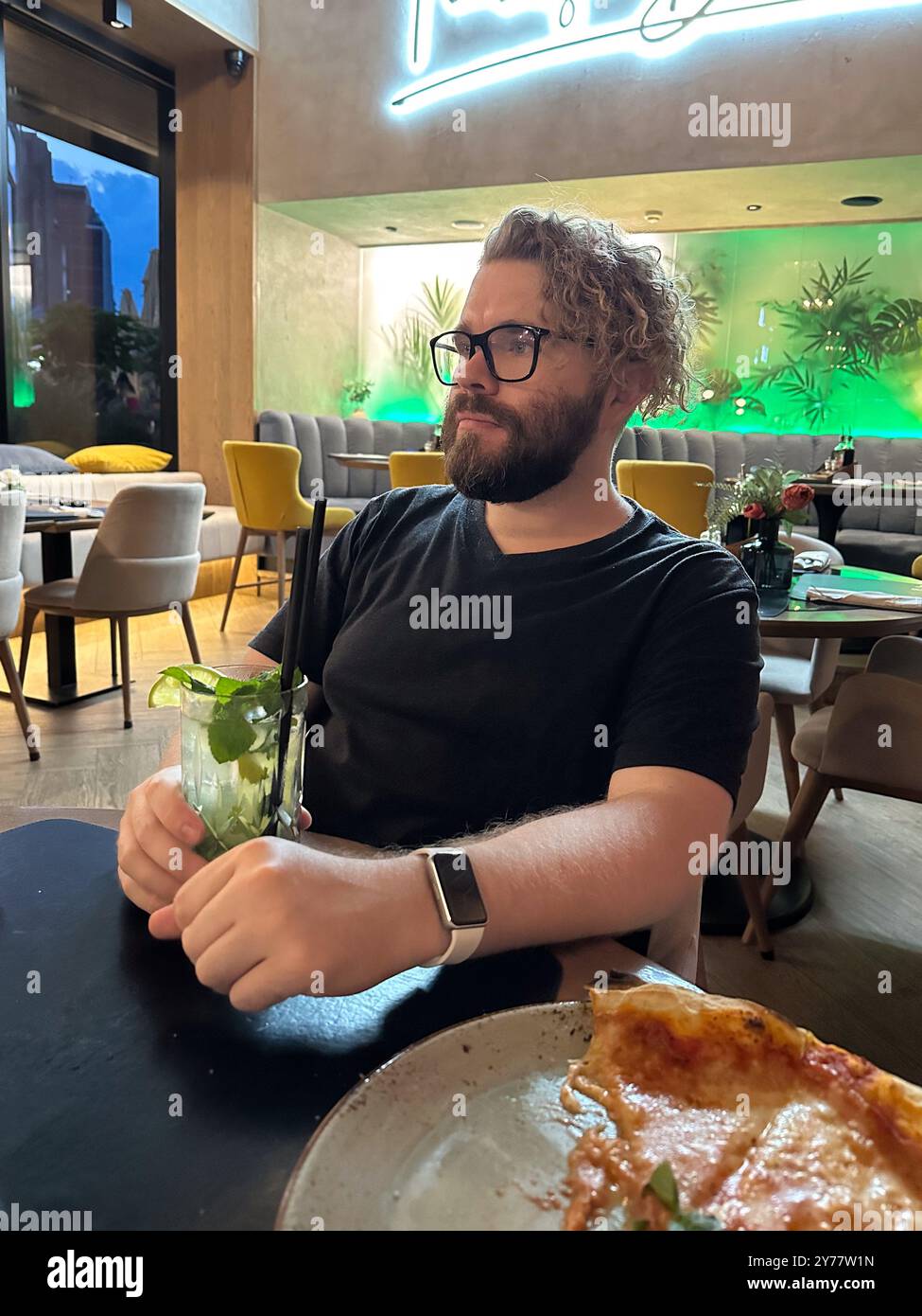 Millennial Curly man mit fröhlichem Ausdruck genießt einen erfrischenden Drink in einem modernen, lebhaften Café-Ambiente. Lebhaftes Dekor im Restaurant mit entspannter Atmosphäre Stockfoto