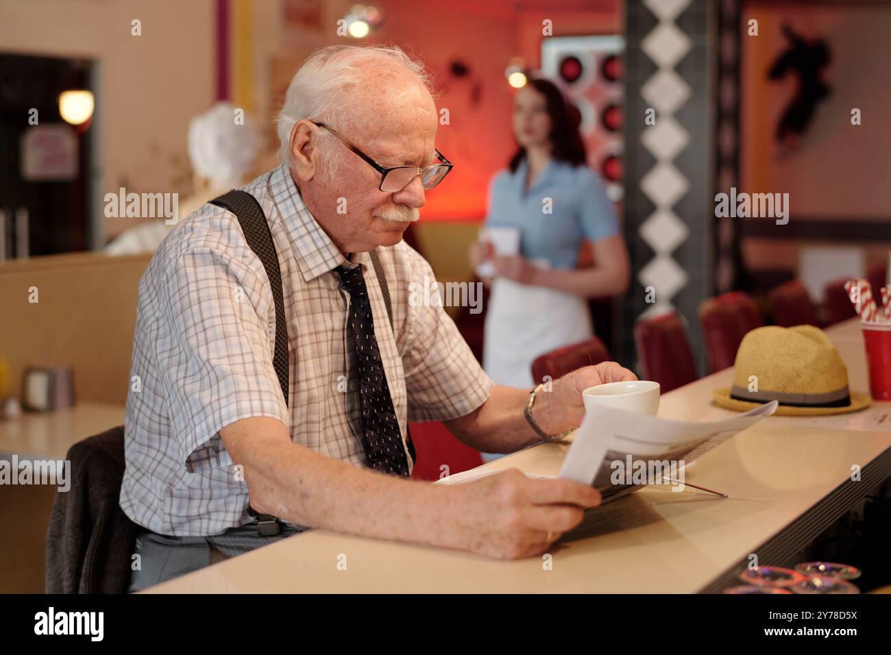 Senior Mann sitzt an der Theke und liest Zeitungen mit Kellnerin im Hintergrund, die einen Kunden bedient. In der Nähe stehen Hocker und Tassen, die eine gemütliche Umgebung schaffen Stockfoto