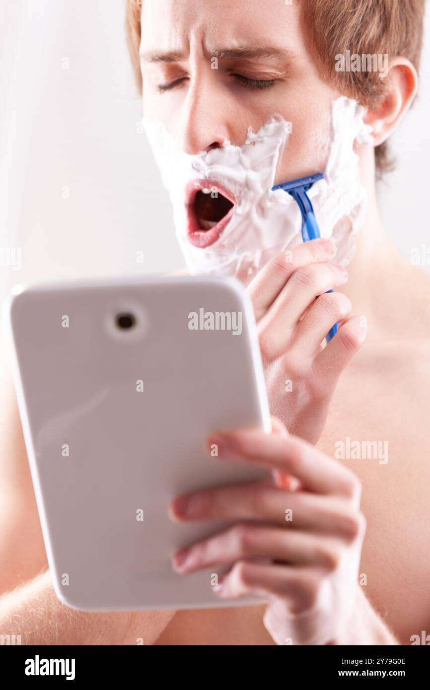 Ein halb wach junger Mann verwendet kreativ ein digitales Tablet als Spiegel während der Rasur und hält ein modernes Morgenritual fest Stockfoto