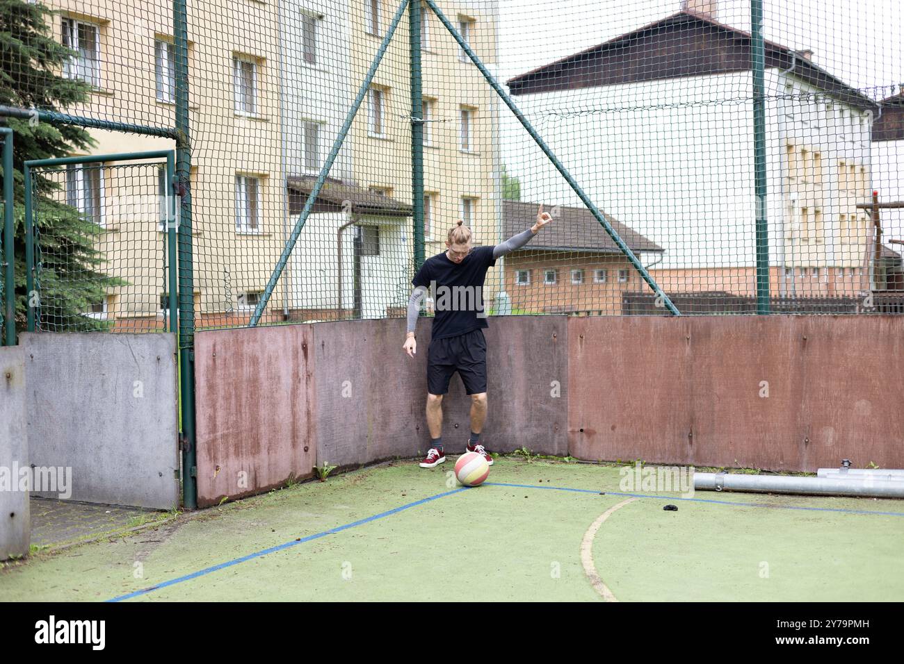 Ein junger Mann steht auf einem Freigelände gegen einen Zaun und signalisiert mit der Hand, während ein Fußball nahe seinem Fuß liegt. Die Szene kombiniert einen Freizeitsport Stockfoto