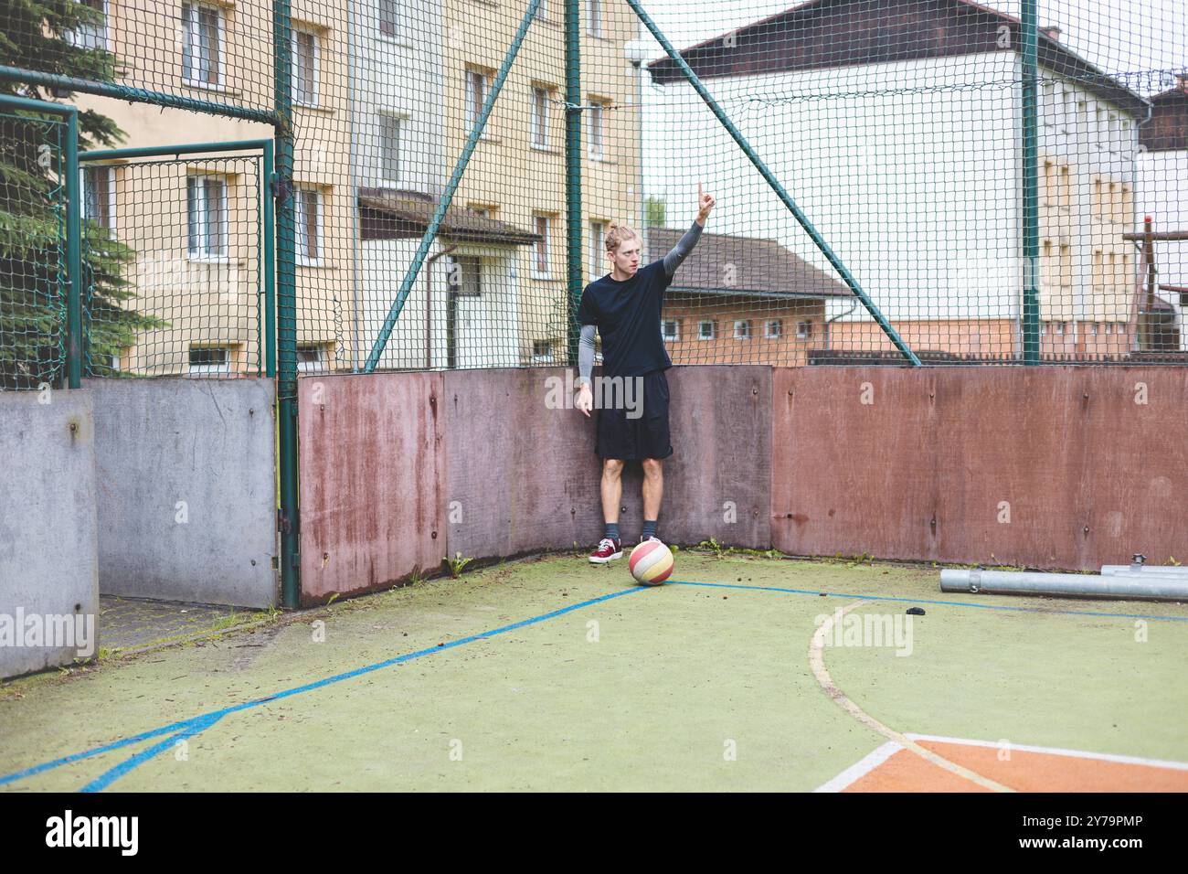 Ein junger Mann steht auf einem Freigelände gegen einen Zaun und signalisiert mit der Hand, während ein Fußball nahe seinem Fuß liegt. Die Szene kombiniert einen Freizeitsport Stockfoto