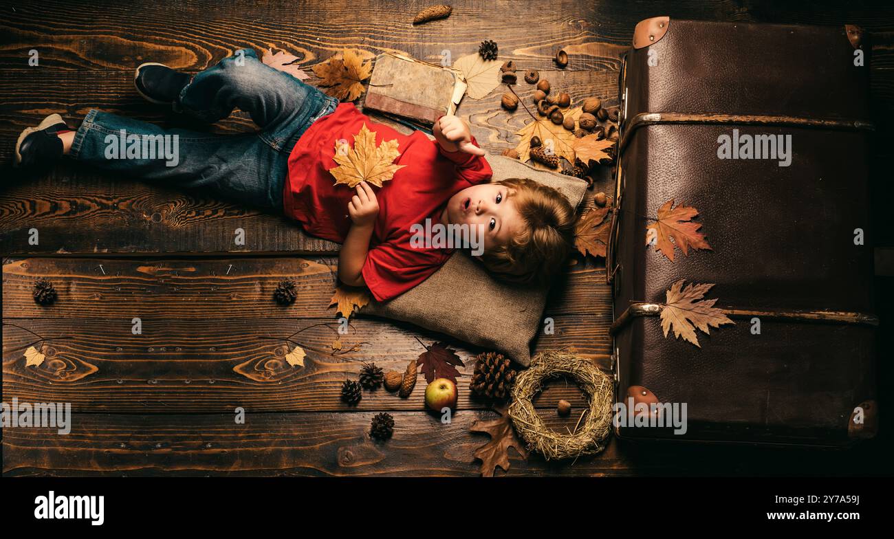 Blonde kleine Junge ruht mit Blatt auf dem Bauch liegt auf Holzboden in Herbstblättern. Der kleine Junge liegt auf einer warmen Decke und träumt von einem warmen Herbst Stockfoto