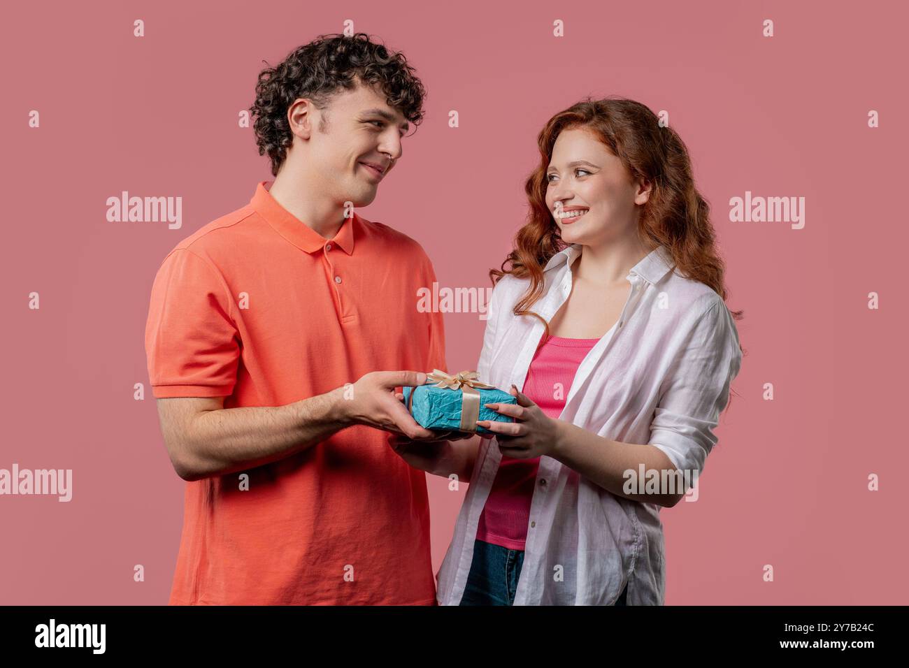 Junges amerikanisches Paar auf rosa Studiohintergrund. Weiße Frau schenkt ihrem Ehemann oder Freund ein Geschenk mit Verbeugung. Geburtstag, Feier, Überraschung, Hochzeitstag Stockfoto