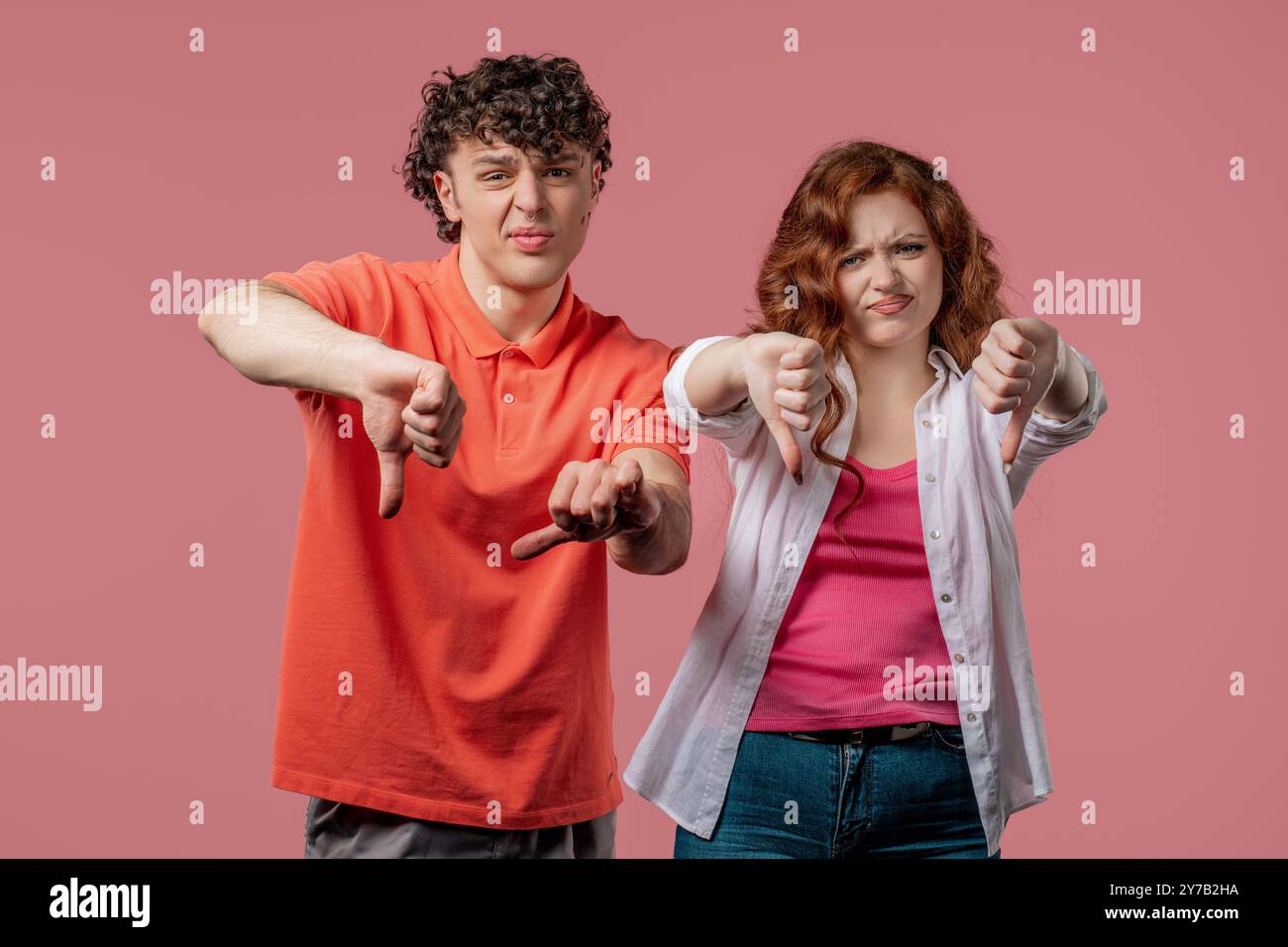 Porträt der unglücklichen europäischen Frau und des Mannes verurteilt mit Zeichen der Abneigung. Ein Paar, das unzufrieden mit der Geste „Daumen nach unten“ im pinken Studio ist Stockfoto