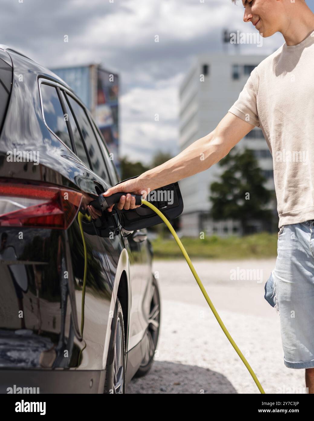 Mann, der eine Steckdose öffnet und ein Ladegerät in ein schwarzes Elektroauto an eine EV-Ladestation in der Stadtstraße stellt Stockfoto