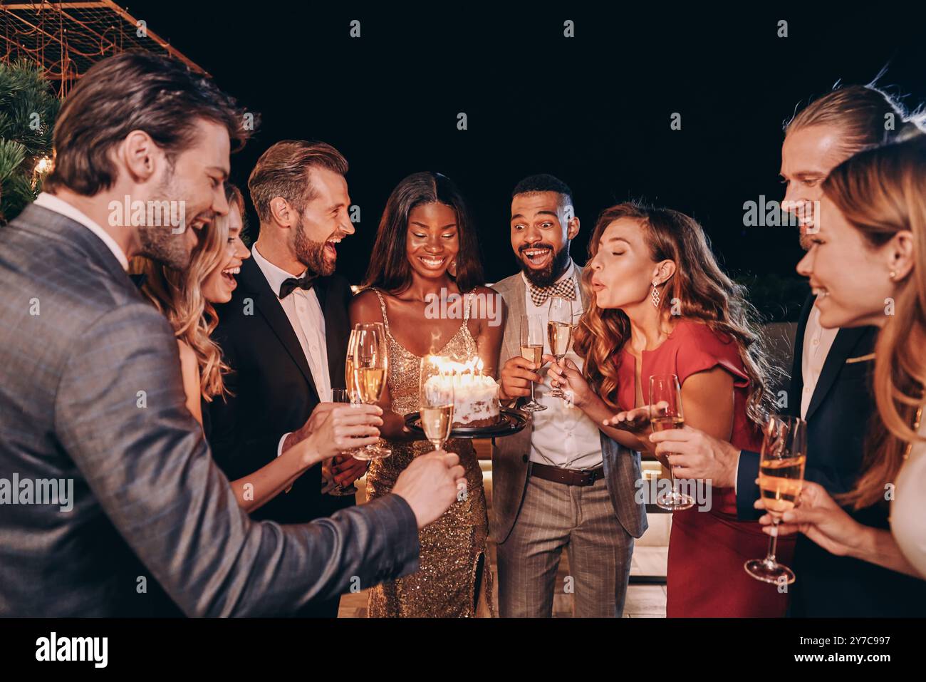 Schöne junge Frau im Abendkleid, die Kerzen auf Kuchen bläst, während sie mit Freunden Geburtstag feiert Stockfoto