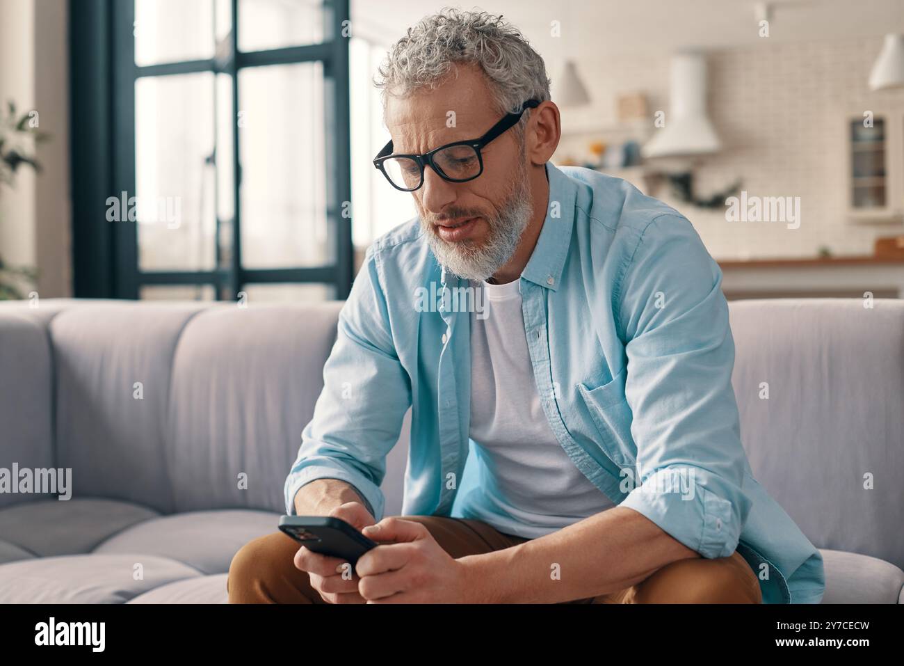 Seniorenmann in lässiger Kleidung und Brille mit Smartphone, während er zu Hause auf dem Sofa sitzt Stockfoto Seniorenmann in lässiger Kleidung und Brille mit Smartphone, während er zu Hause auf dem Sofa sitzt Stockfoto