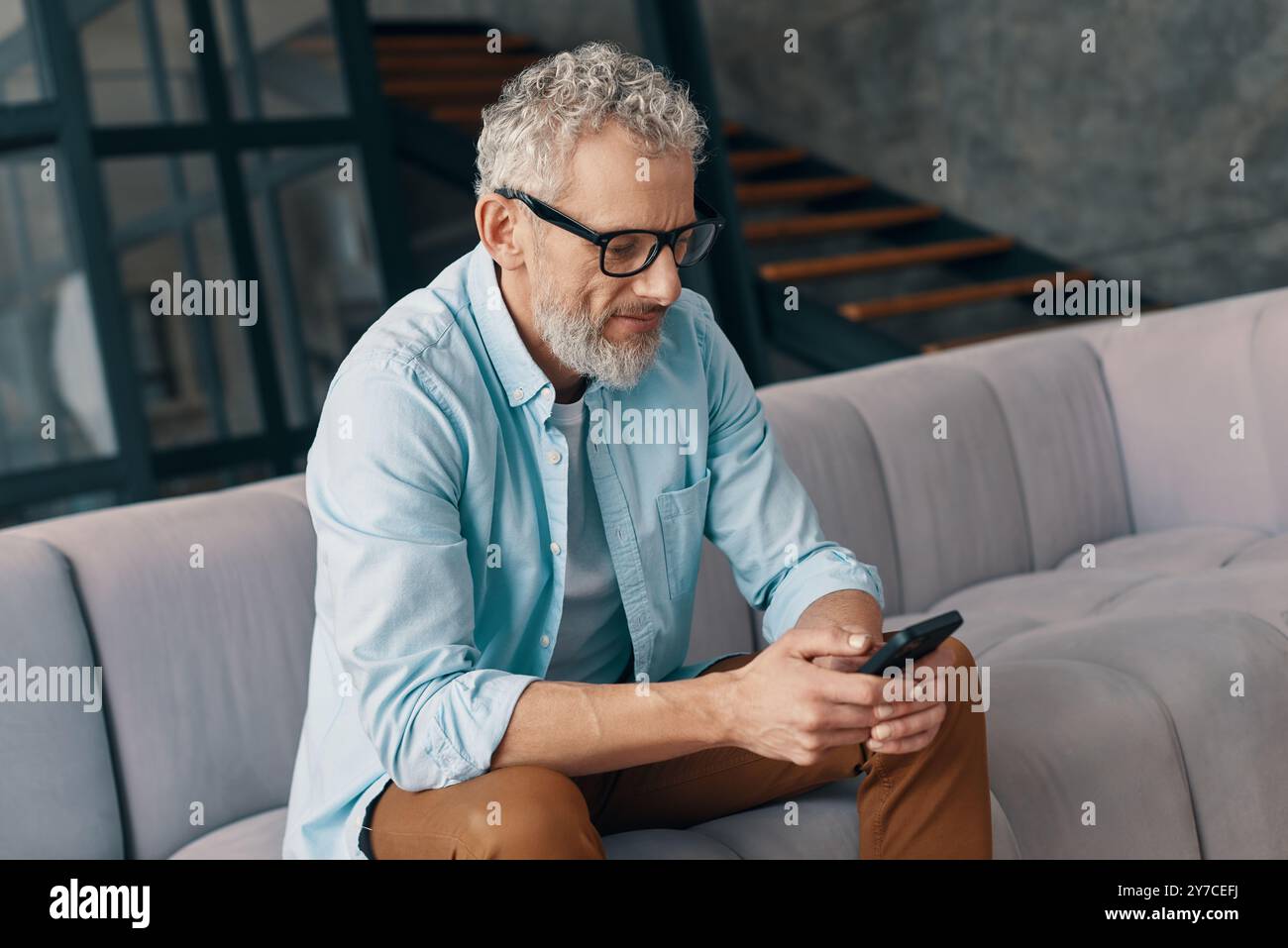 Seniorenmann in lässiger Kleidung und Brille mit Smartphone, während er zu Hause auf dem Sofa sitzt Stockfoto Seniorenmann in lässiger Kleidung und Brille mit Smartphone, während er zu Hause auf dem Sofa sitzt Stockfoto