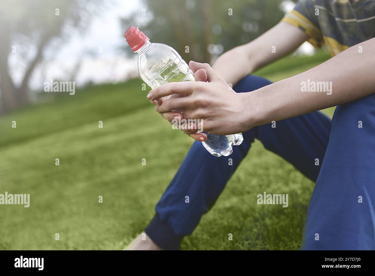 Jungen kaukasischen Mann runner entspannende Holding Trinkwasser Flasche und sitzen auf dem Rasen im Park im Freien nach Sport am Morgen Zeit, Exercis Stockfoto