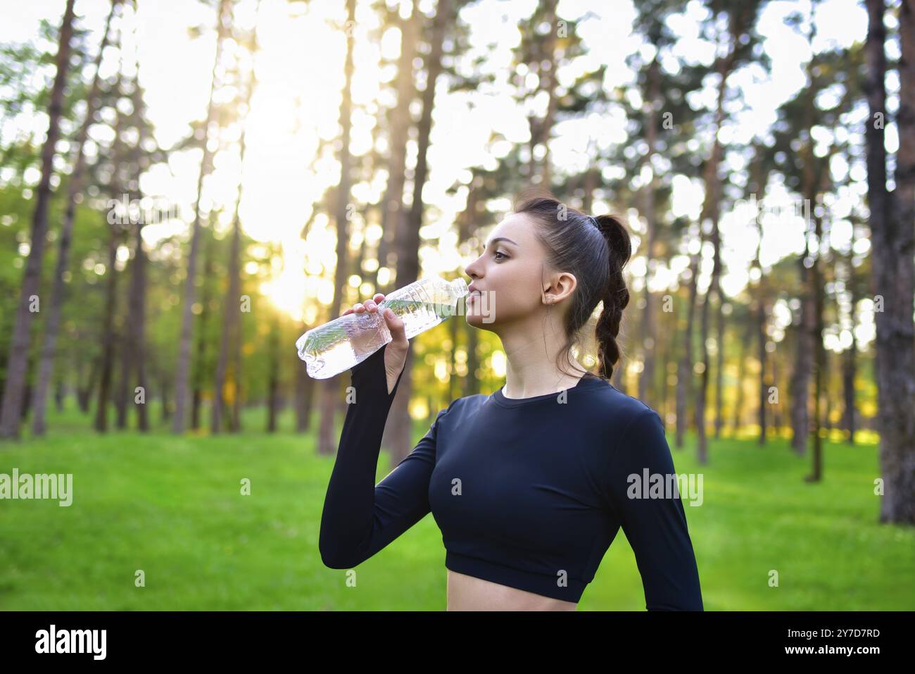 Eine Frau in einem sportlichen schwarzen Oberteil hält nach einem Sport im Wald eine Wasserflasche hoch, um Wasser zu trinken Stockfoto