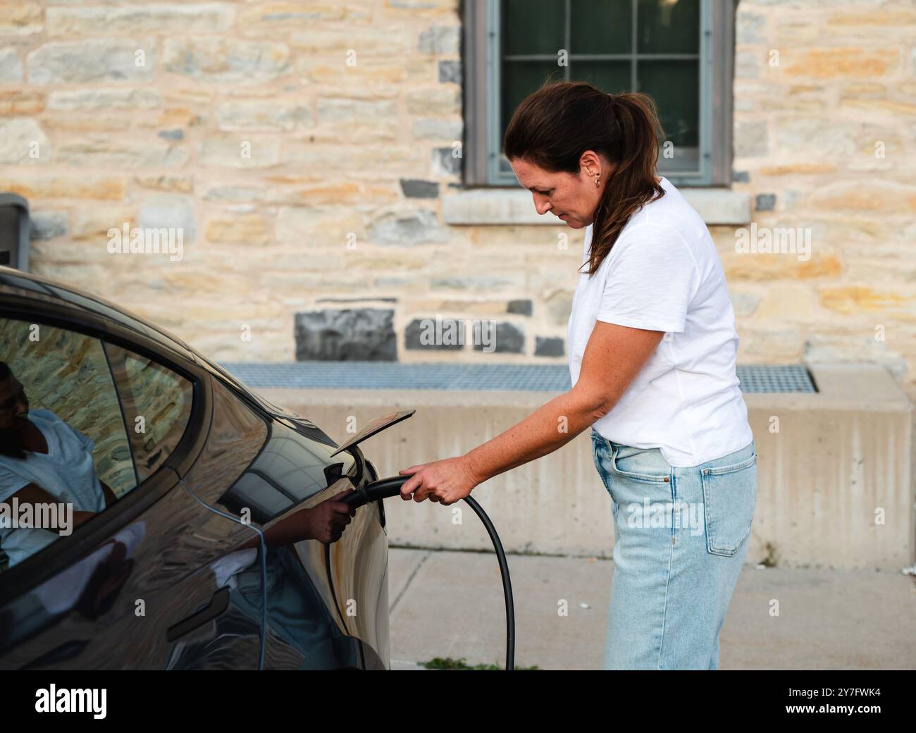 Frau, die draußen ein EV-Ladegerät an ein Elektrofahrzeug angeschlossen hat. Stockfoto