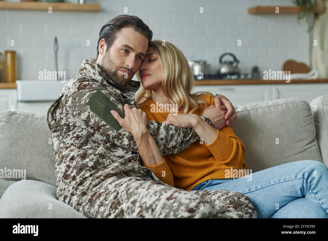 Trauriger junger Mann in militärischer Form, der seine Frau umarmt, während er zu Hause auf der Couch sitzt Stockfoto