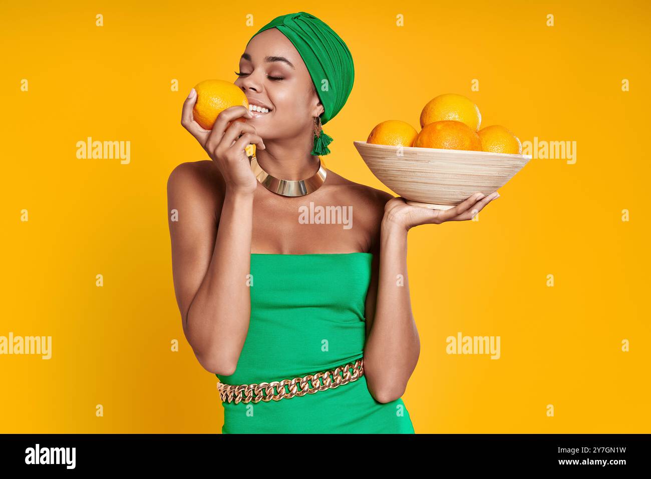 Schöne afrikanische Frau in traditionellen Kopfbedeckungen, die Orangen riecht und vor gelbem Hintergrund lächelt Stockfoto