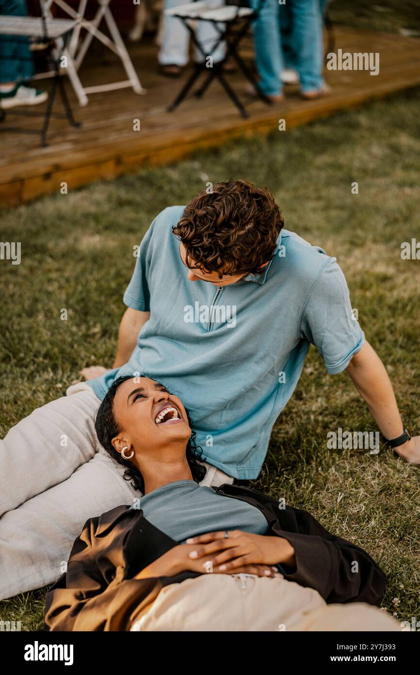 Hochwinkelansicht einer glücklichen Frau, die auf dem Schoß eines männlichen Freundes liegt, der auf Gras im Hinterhof sitzt Stockfoto