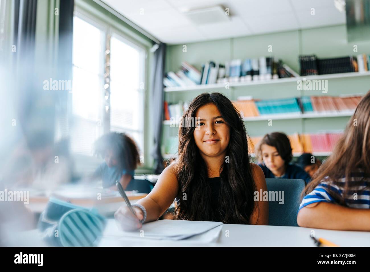 Porträt eines lächelnden Mädchens mit langen Haaren, das in der Grundschule im Klassenzimmer sitzt Stockfoto