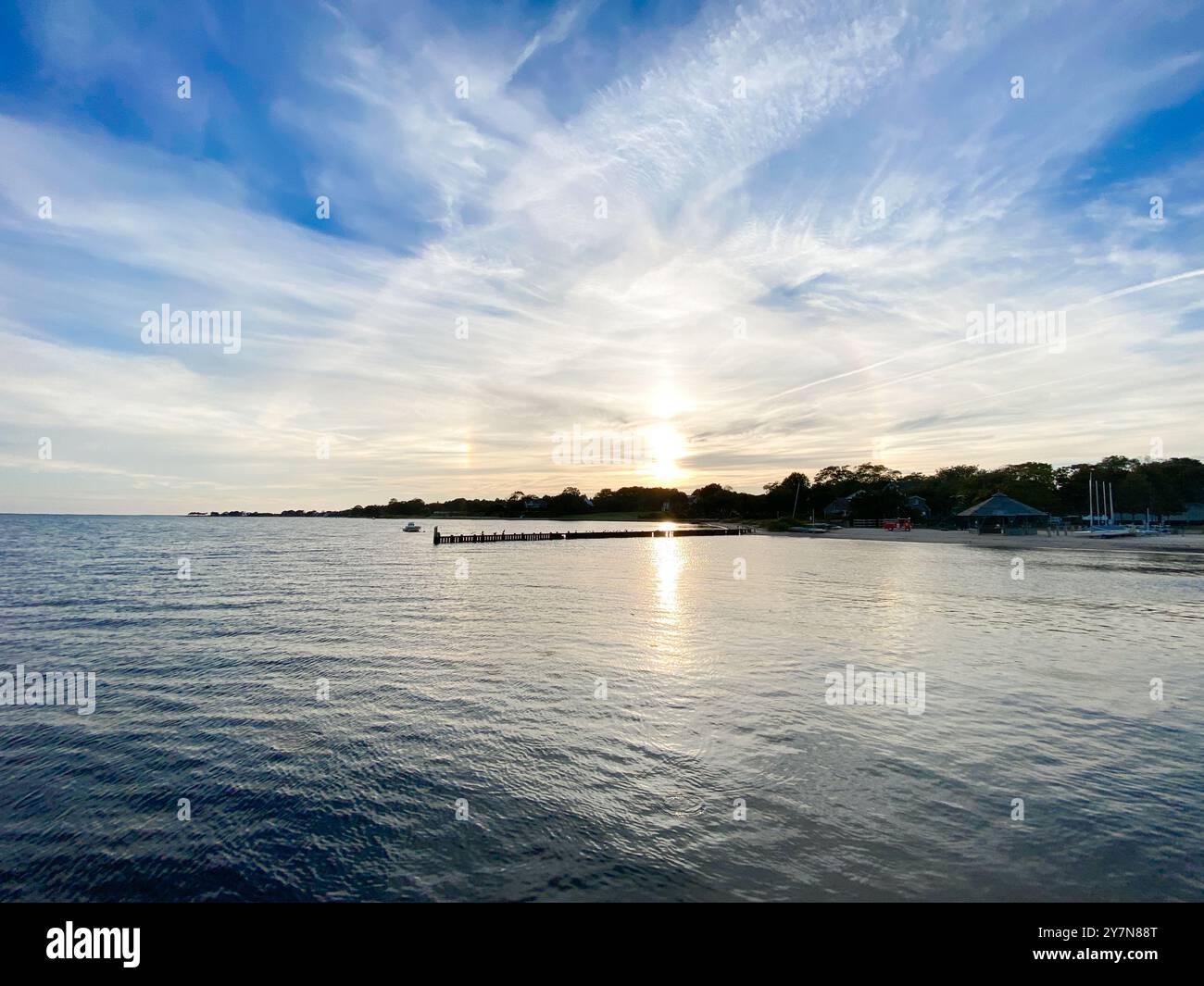 Ruhige Bucht mit Wasser, am Rand des Wassers. Cirrostratus-Wolken am blauen Himmel und goldene Sonne, umringt von einem „Halo“ oder Ring der Lichtbrechung auf den Wolken. Stockfoto
