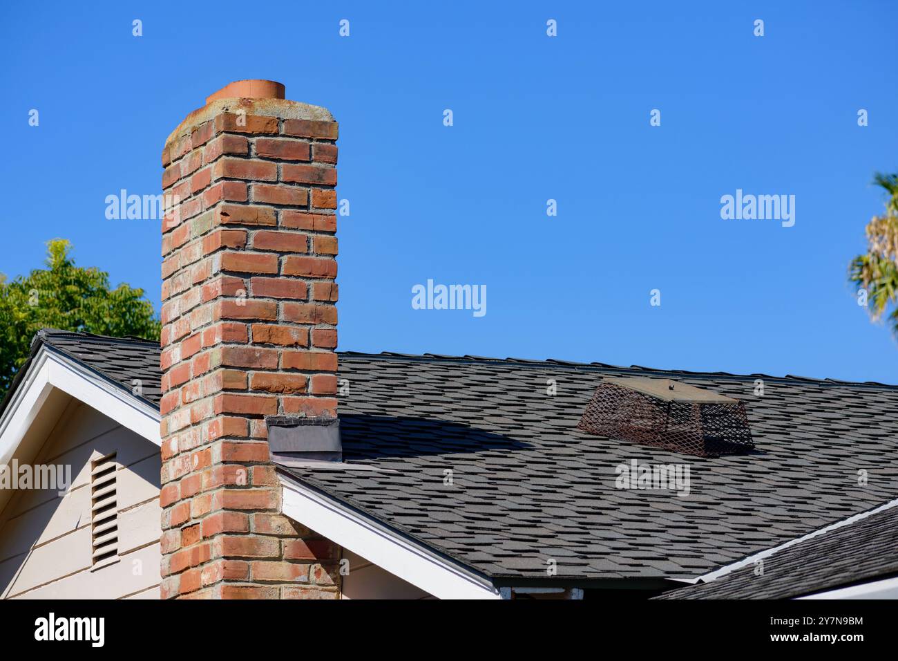 Ein Wohndach mit einem roten Ziegelkamin, dessen Schornsteinkappe auf den schwarzen Schindeln verlagert liegt. Stockfoto