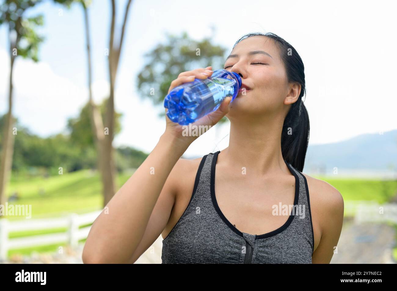 Eine schöne, fitte asiatische Frau in Sportbekleidung trinkt aus einer Wasserflasche und löscht ihren Durst nach dem Joggen in einem grünen Park an einem hellen Tag. Sommer-Akti Stockfoto