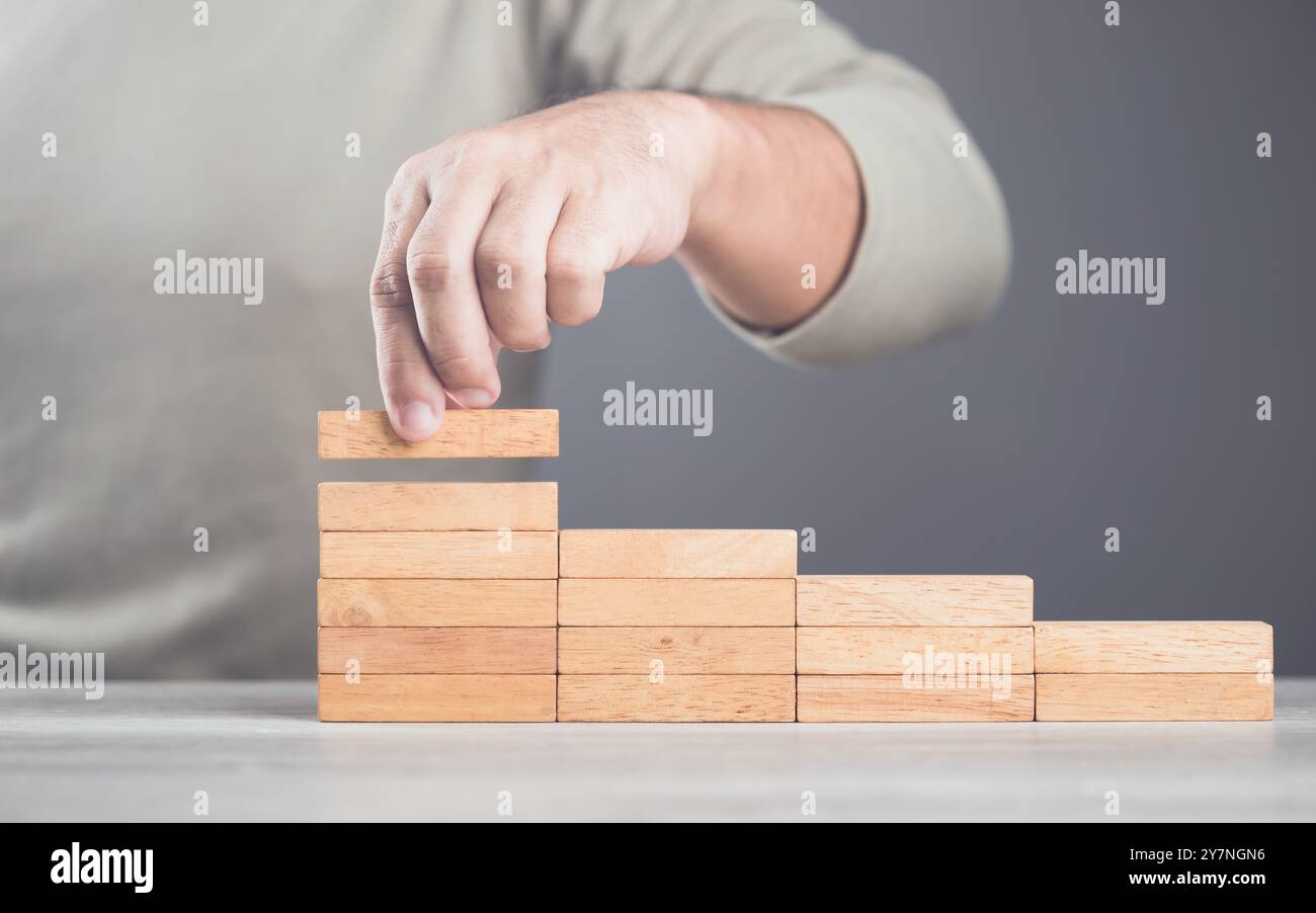 Die Hand eines Geschäftsmannes stapelt braune Holzblöcke von niedrig bis hoch und symbolisiert damit Geschäftsstrategie und Wachstum. Das Bild zeigt den Schritt des erfolgreichen Aufbaus Stockfoto