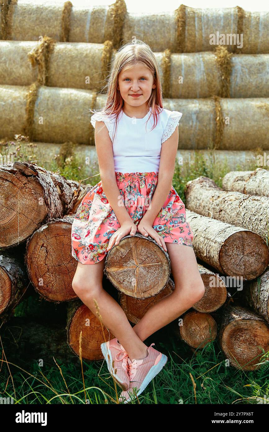 Ein junges Mädchen mit langen, lockeren Haaren sitzt in einem rosafarbenen Kleid auf einem Baumstamm und ihre Beine hängen draußen an einem warmen Sommertag Stockfoto