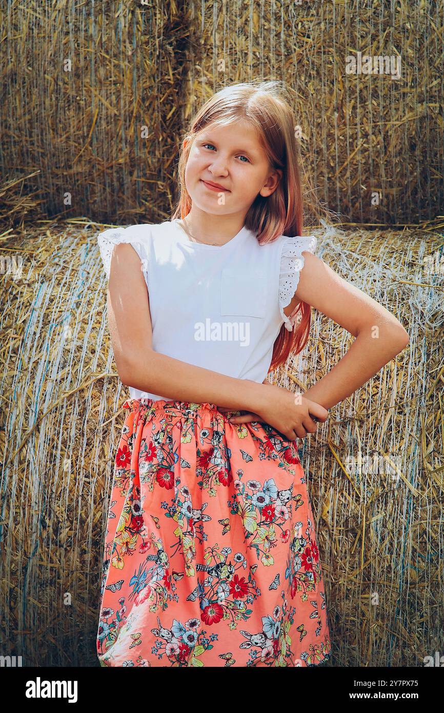 Ein blondes Mädchen mit langen Haaren und einem Kleid steht an einem sonnigen heißen Tag neben Strohballen Stockfoto