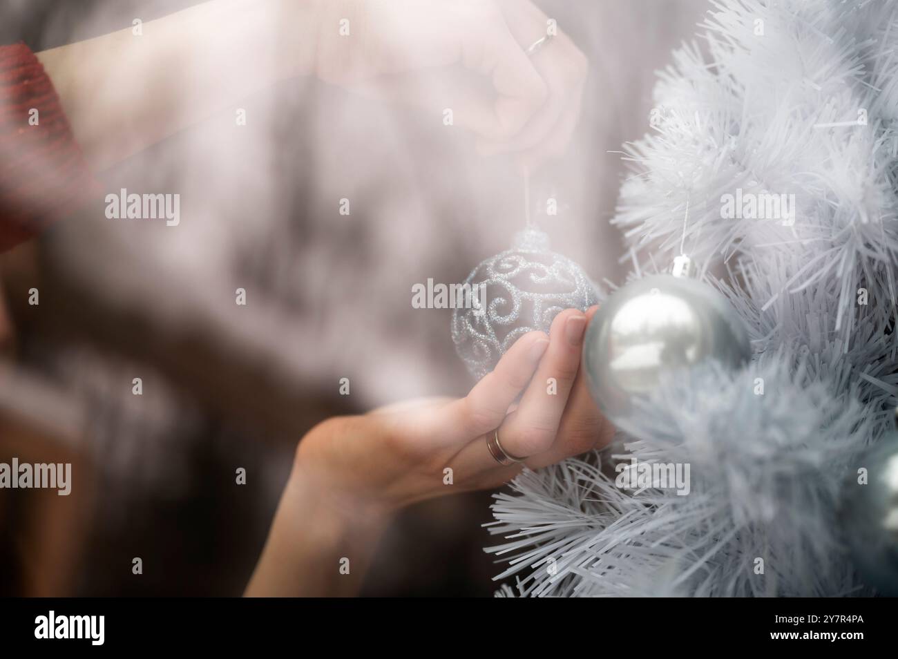 Blick durch das Fenster der weiblichen Hand, die weißen Weihnachtsbaum mit glänzender lacey-blauer Weihnachtskugel dekoriert. Mit Reflexion des Außen in der gla Stockfoto