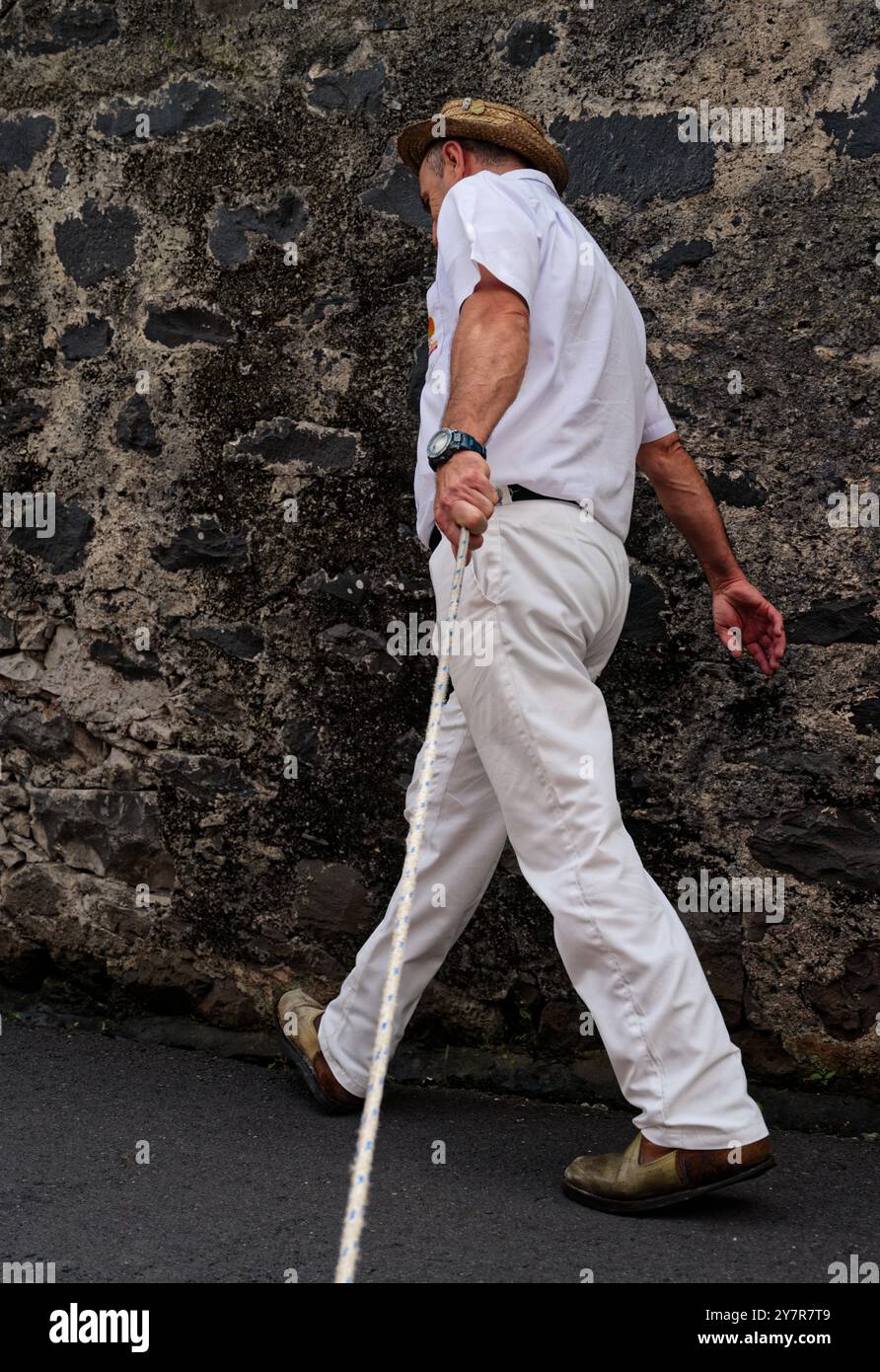 Ein Karreiro hält ein Seil, während er an einer Steinmauer entlang läuft und sich auf die nächste Rodelfahrt mit der Korbbahn vorbereitet Stockfoto