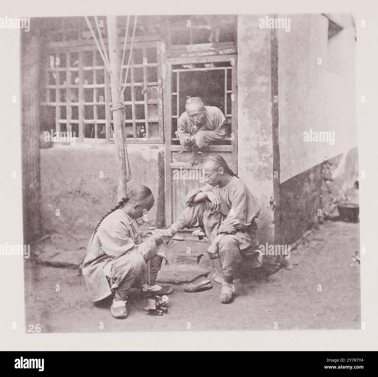 Vintage-Foto eines fahrenden Chiropodisten, der auf einem Mais am Fuß seines Patienten operiert. Peking, Nord-Zhili (Provinz Pei-chih-li oder Peking), China. Das Motiv der Illustration ist ein reisender Chiropodist, der auf einem Mais operiert und die Zehennägel eines Kunden anzieht; während ein zweiter Patient ruhig wartet, bis er selbst an der Reihe ist und die Pfeife des Friedens aus einem kaputten Fenster raucht. Stockfoto