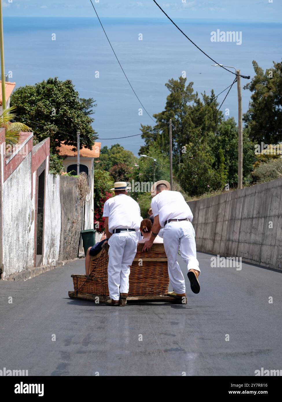 Carreiros schiebt eine Korbrodelbahn eine ruhige Straße hinunter, während sich der atlantik in Funchal ausdehnt Stockfoto