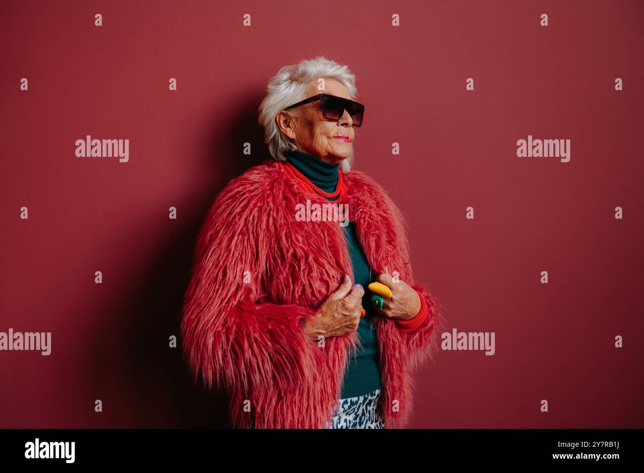 Modische ältere Frau in stilvoller Brille und trendiger Kleidung, die auf rotem Hintergrund steht Stockfoto