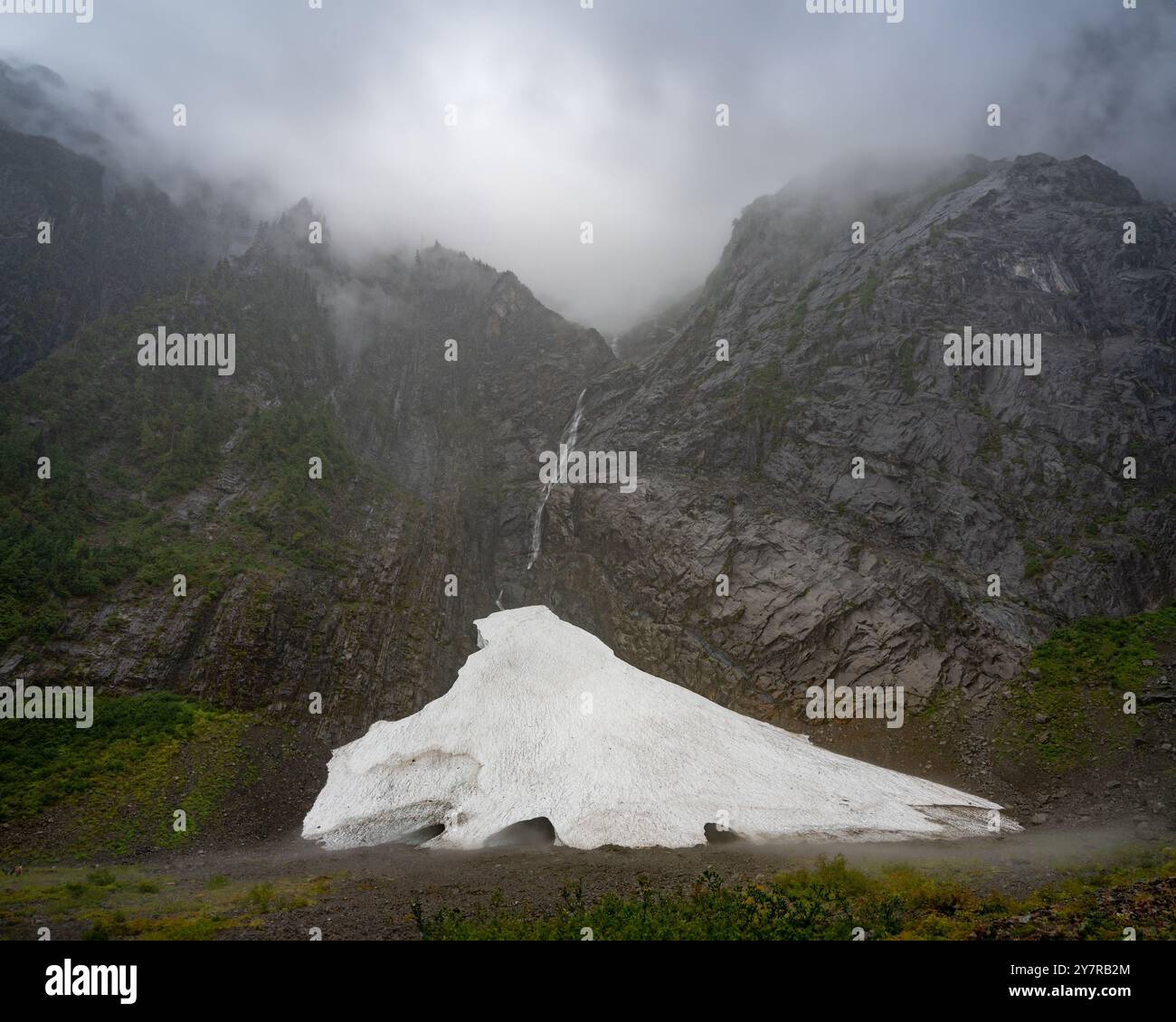 Eishöhle am Fuße eines nebeligen Berges mit einem Wasserfall, der durch felsige Klippen fließt, umgeben von Nebel und Wolken in einer abgelegenen Wildnis. Stockfoto