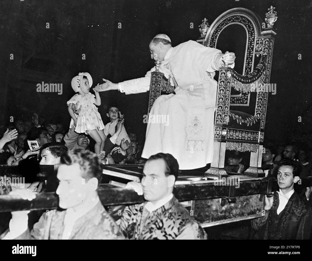 DER PAPST SEGNET KINDER viele Kinder waren in der großen Menge von Pilgern, die der Papst diese Woche in der Audienz des Petersdoms empfangen hat, bevor er Rom zu seinem Sommerpalast nach Castelgandolfo verlässt. Das Foto zeigt Papst Pius XII. Lehnt sich von seinem Stuhl, um das Gesicht eines kleinen Kindes zu berühren, das ihn schüchtern ansieht, als es aus der Menge gehoben wird. 15. Juli 1953 Stockfoto