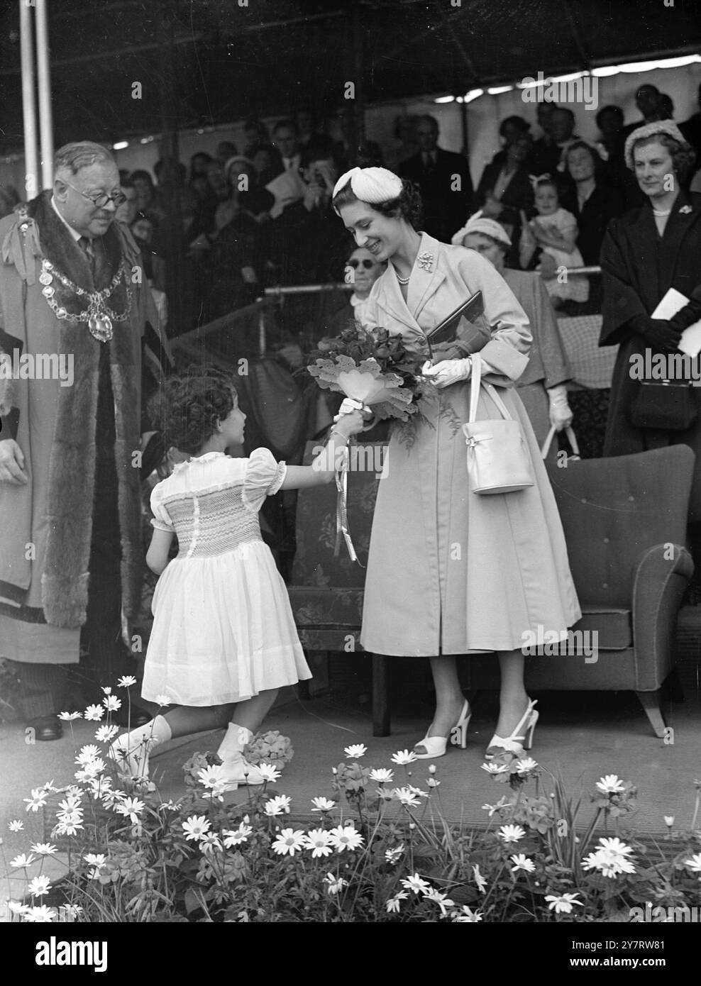 DIE PRINZESSIN BEIM WETTBEWERB 6,53 UHR. Prinzessin Margaret flog gestern nach Bridport, Devon, um an der Eröffnungsaufführung eines Festwettbewerbs teilzunehmen, der den 700. Jahrestag der Verleihung der Königlichen Charta der Stadt feierte. Das Foto zeigt den Bürgermeister von Bridport, Alderman H. R. C. Palmer, sieht mit Zustimmung zu, wie seine 9-jährige Nichte einen klugen Knick macht, während sie Prinzessin Margaret mit einem Blumenstrauß präsentiert. 25. Juni 1953 Stockfoto
