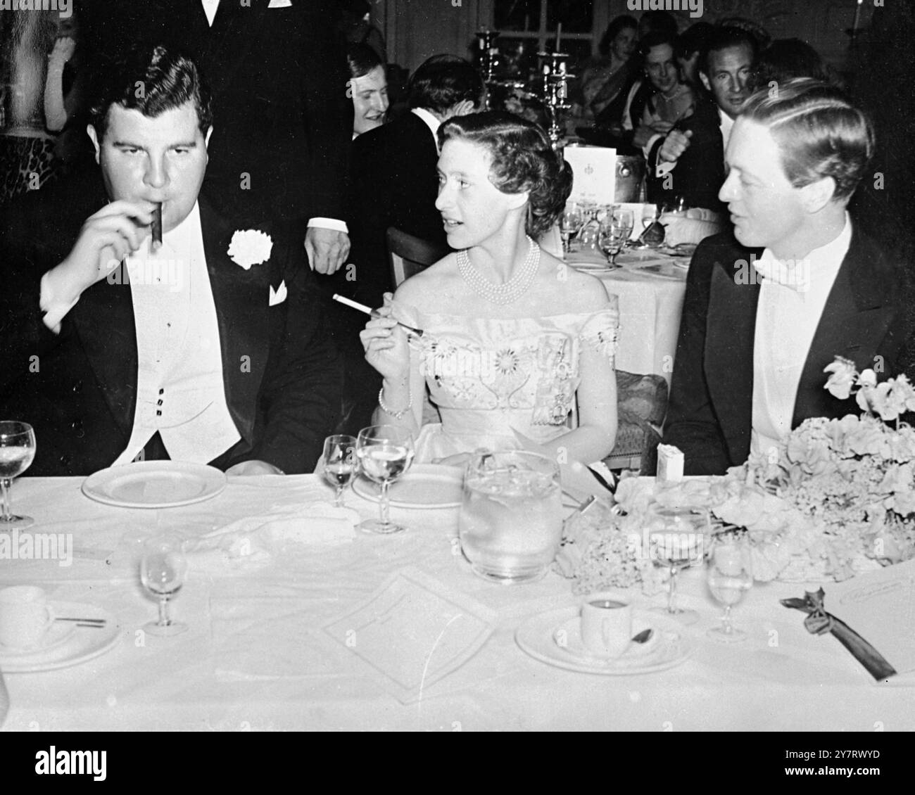 Prinzessin im Ball Prinzessin Margaret besuchte gestern Abend einen Wohltätigkeitsball zugunsten der Dockland Siedlung im Savoy Hotel, London Foto zeigt: Prinzessin Margaret genießt zwischen den Tänzen eine Zigarette. Sie sitzt zwischen zwei ihrer Partner, Jeremy Tree (links) und dem Marquess of Blandford am 27. Mai 1953 Stockfoto