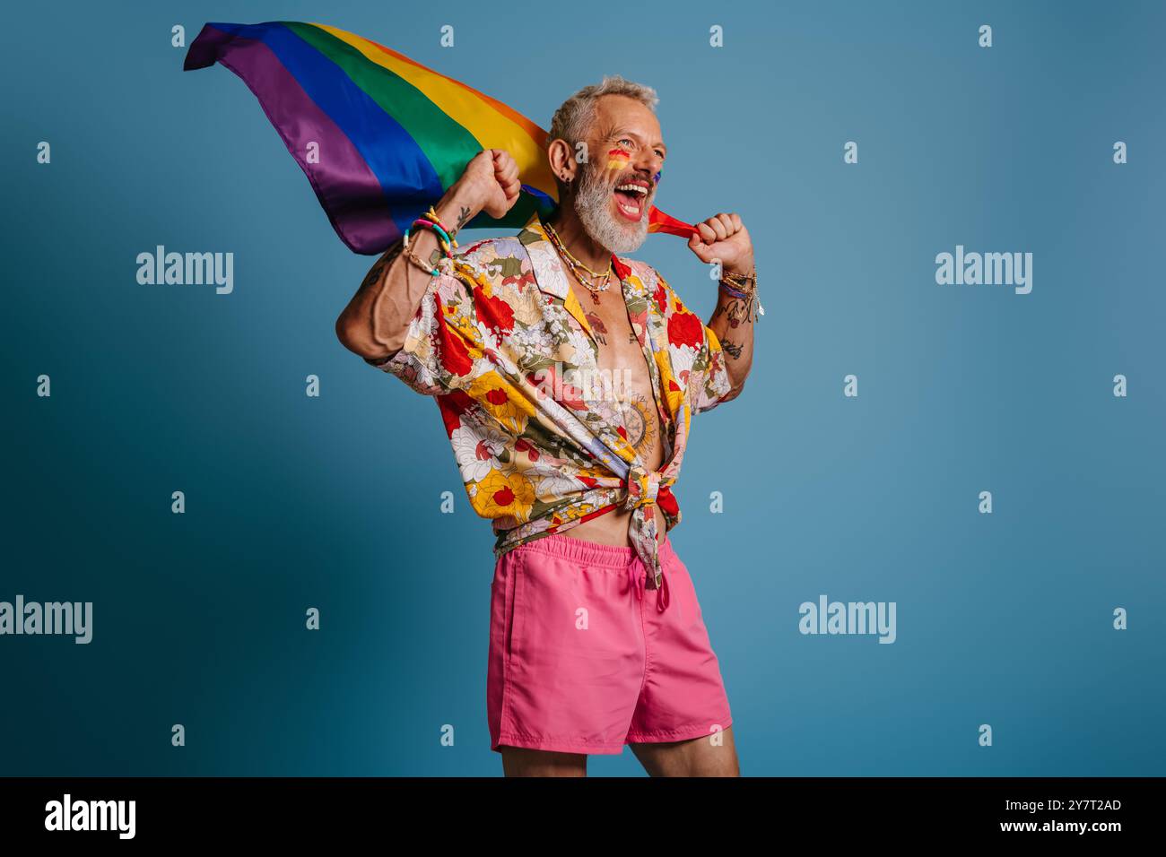 Fröhlicher reifer schwuler Mann mit Regenbogenfahne und Lächeln, während er vor blauem Hintergrund steht Stockfoto