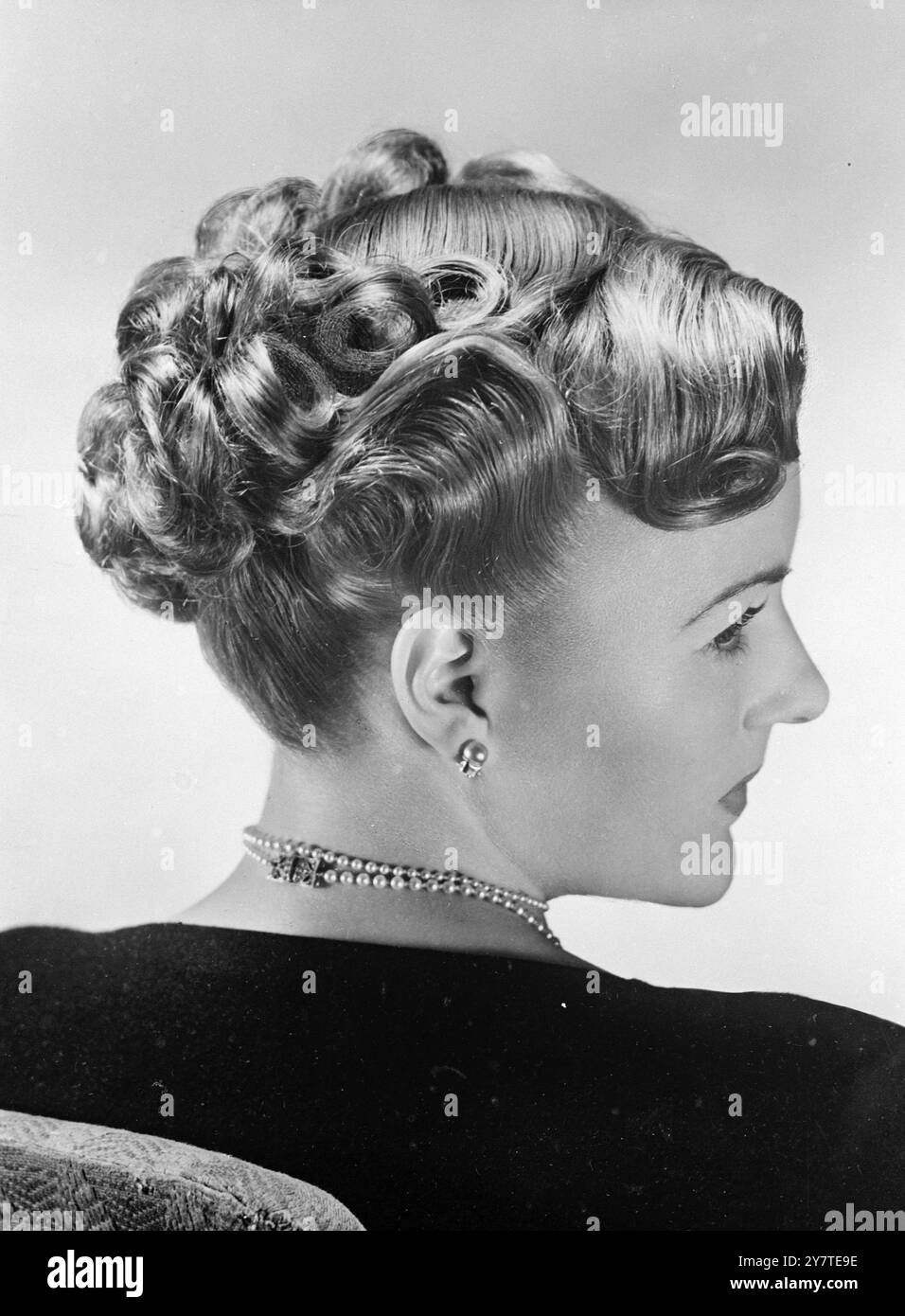 Dieser charmante feminine Stil wurde von Toni für mittellanges Haar entworfen. Die Lockenanordnung des Pferdeschuhs wird erreicht, indem die Enden an der Rückseite und Seite sanft durchgedrückt werden und dann das Haar in Pincurls von der Nackenbasis bis zur Kopfkrone in ordentlichen Schichten aufgetragen wird. Das vordere Haar wird seitlich zu einem kurzen Knall gebürstet. Der Stil ist ideal für Frauen, die ihr Haar lang halten und gleichzeitig modisch bleiben möchten. Stockfoto