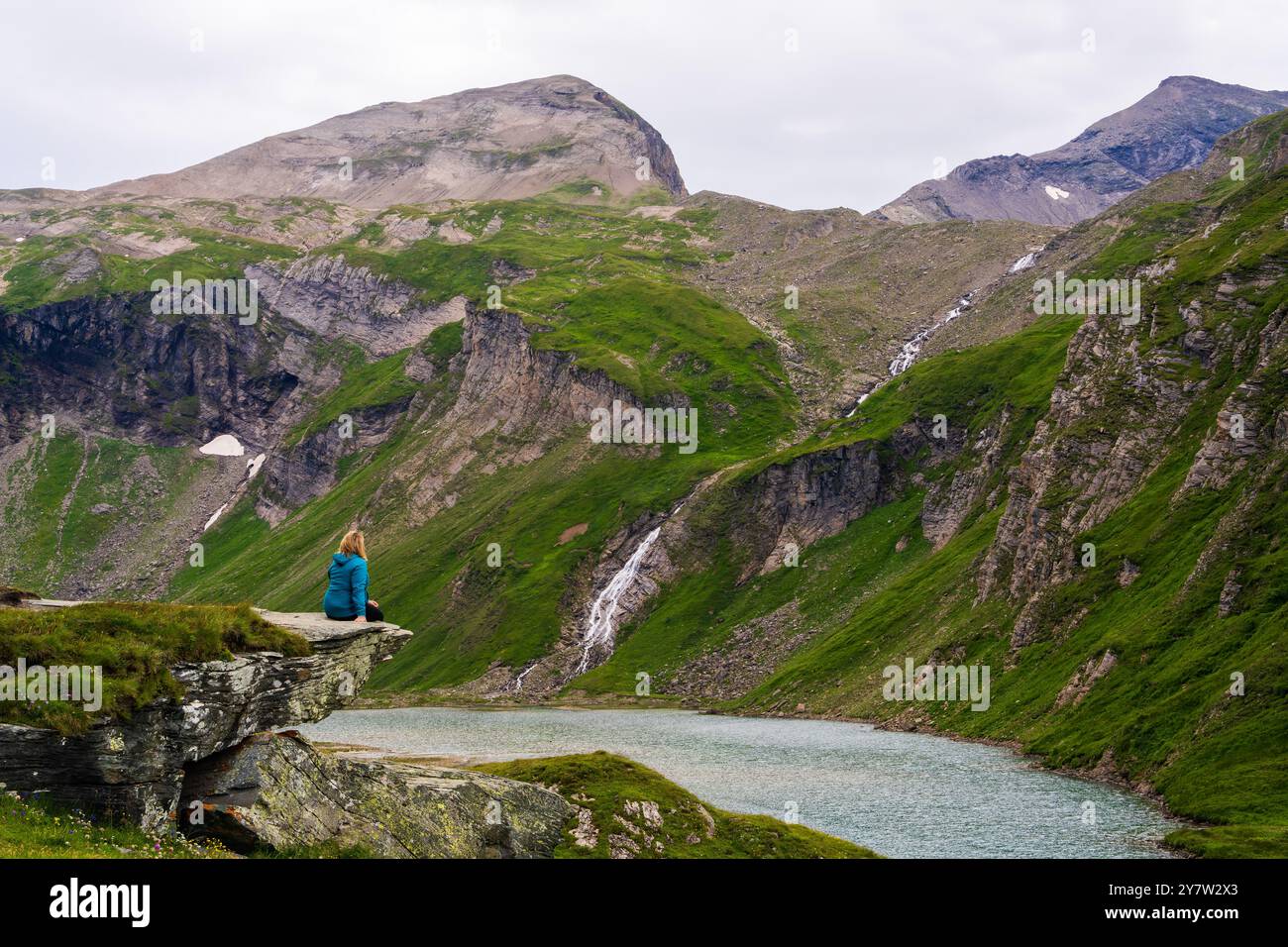 Frau sitzt auf einem großen Felsen, Blick über den blauen Bergsee, umgeben von Bergen, die von grüner Vegetation bedeckt sind. Kaukasische Frau, die nachdenklich den Kampf genießt Stockfoto