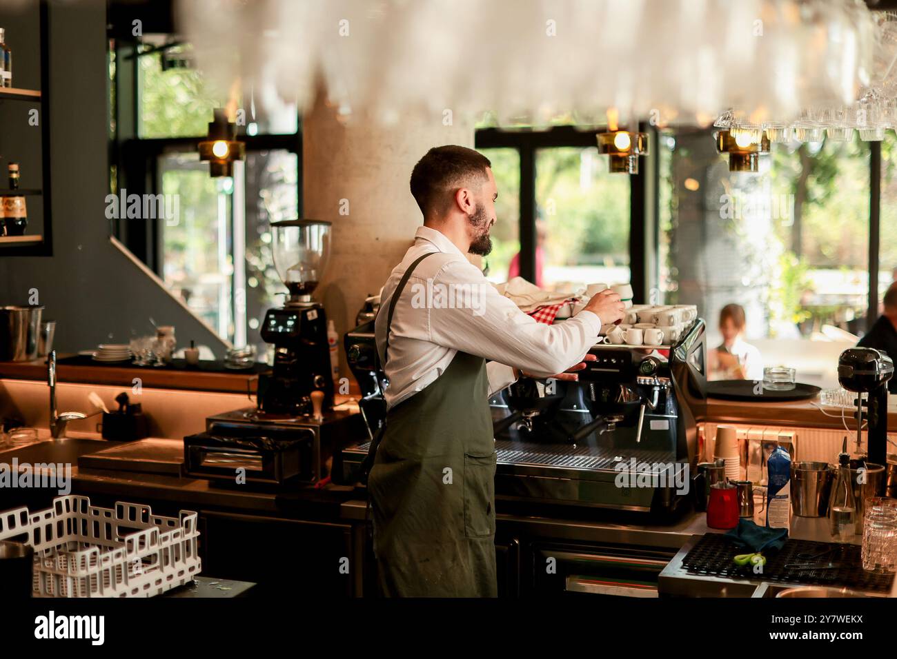 Ein engagierter Barista bereitet Espresso fachmännisch zu und bietet eine lebhafte Atmosphäre voller Sonnenlicht, Pflanzen und zufriedener Gäste, die ihren genießen Stockfoto