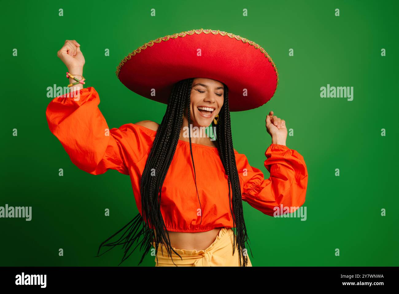 Schöne junge mexikanische Frau in Sombrero, die glücklich aussieht, während sie vor grünem Hintergrund tanzt Stockfoto