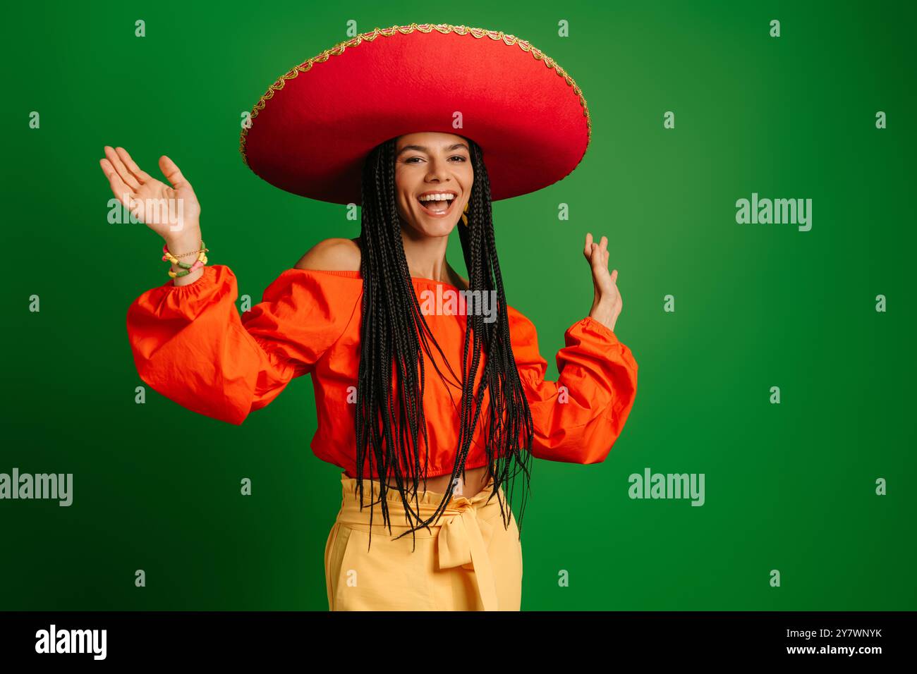 Attraktive junge mexikanische Frau in Sombrero, die glücklich aussieht, während sie vor grünem Hintergrund tanzt Stockfoto