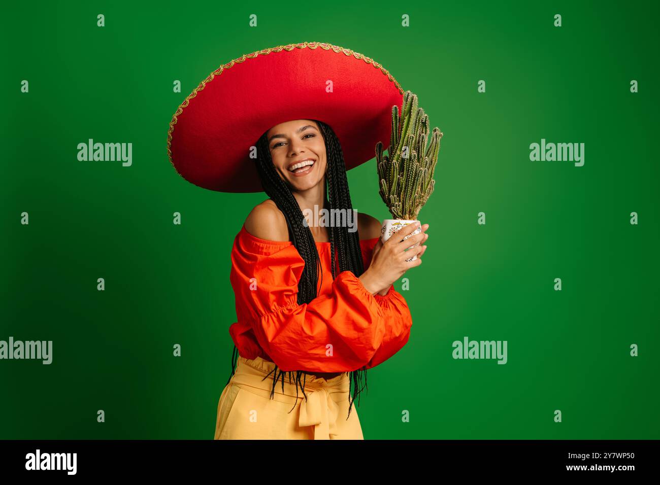 Schöne junge mexikanische Frau in Sombrero, die Kakteen hält und vor grünem Hintergrund lächelt Stockfoto