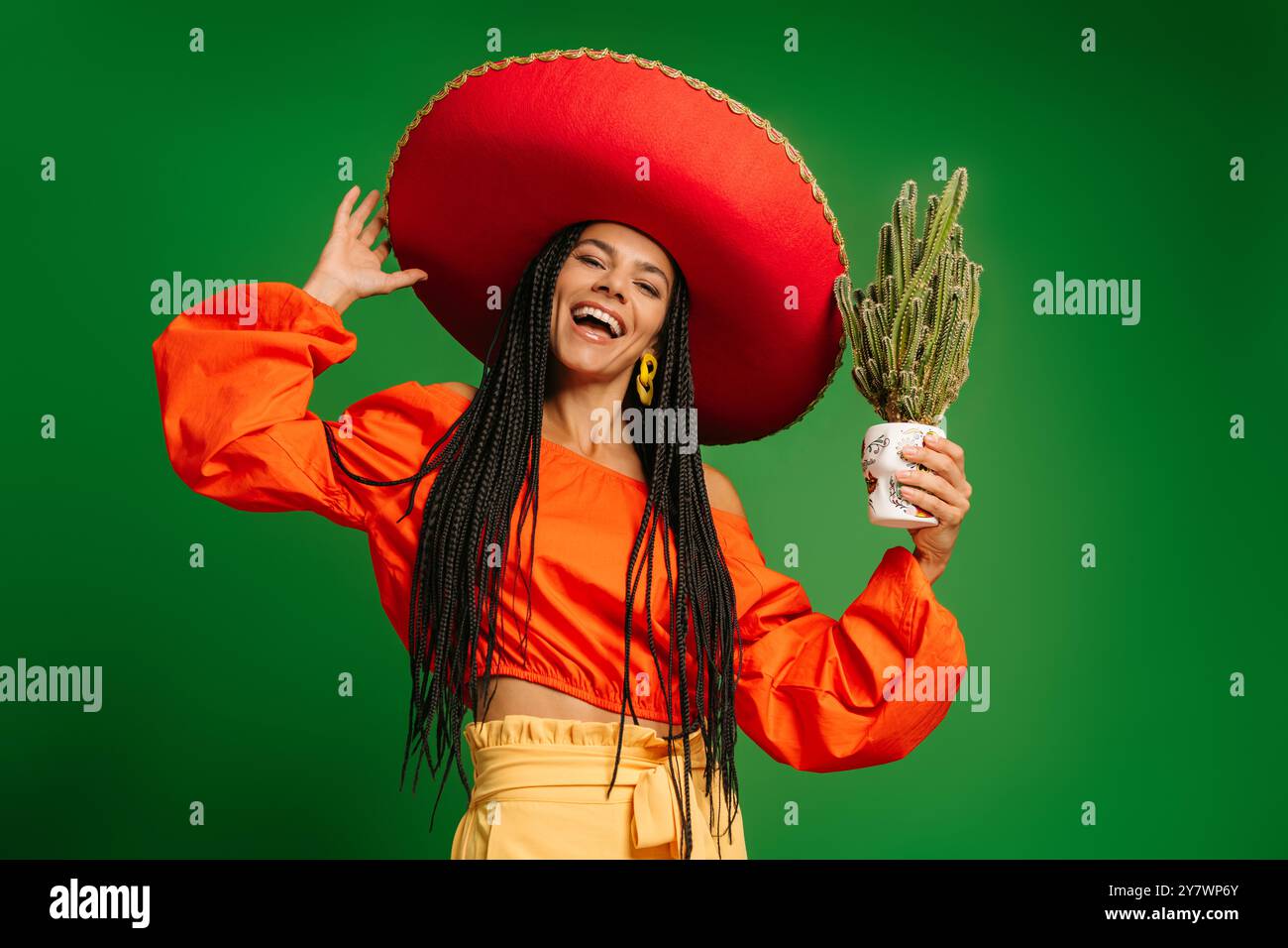 Attraktive junge mexikanische Frau in Sombrero, die Kakteen hält und vor grünem Hintergrund lächelt Stockfoto