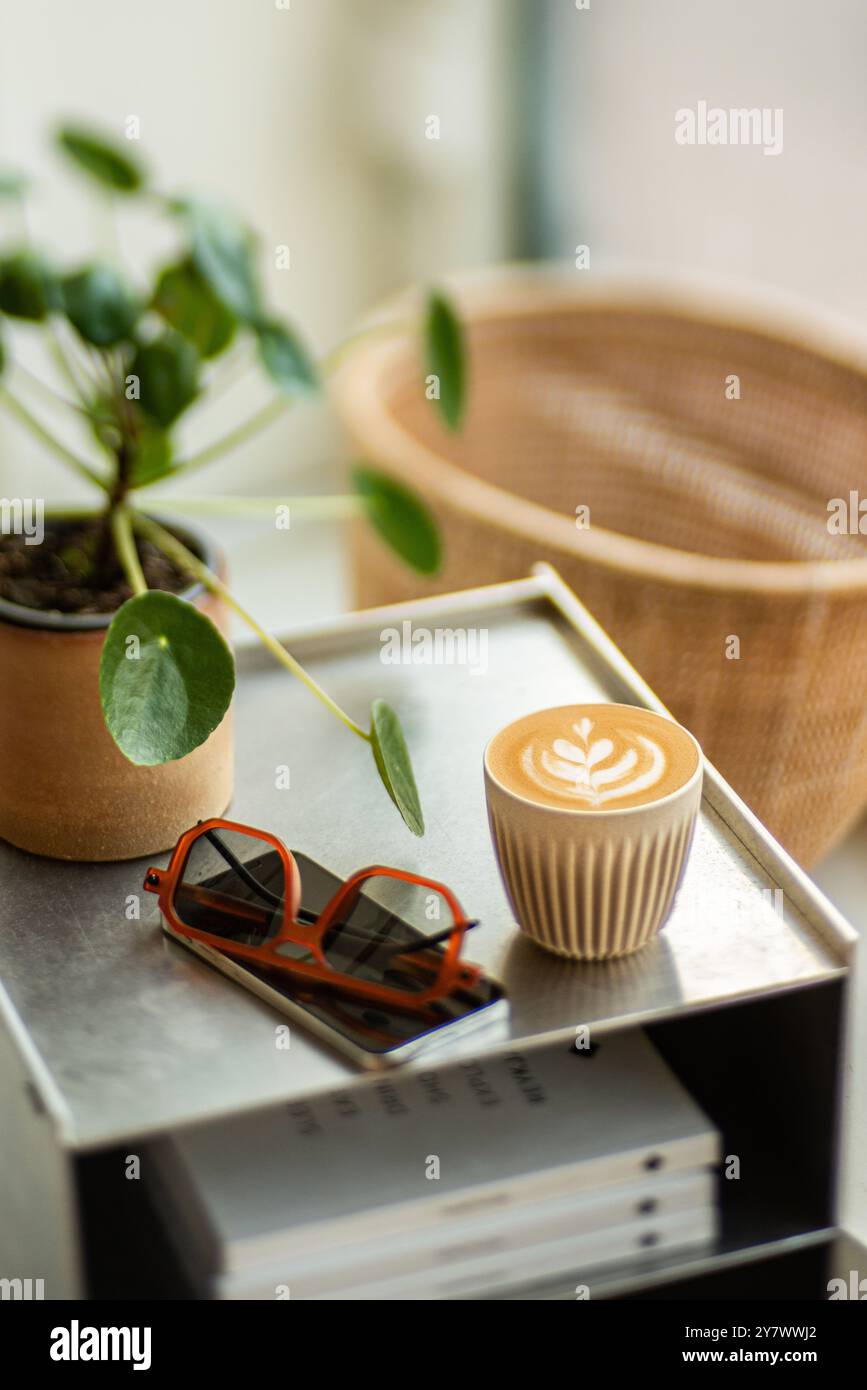Tasse Cappuccino mit wunderschöner Latte Art, auf einem Metall-Nachttisch, umgeben von Pflanzen und Büchern. Stockfoto