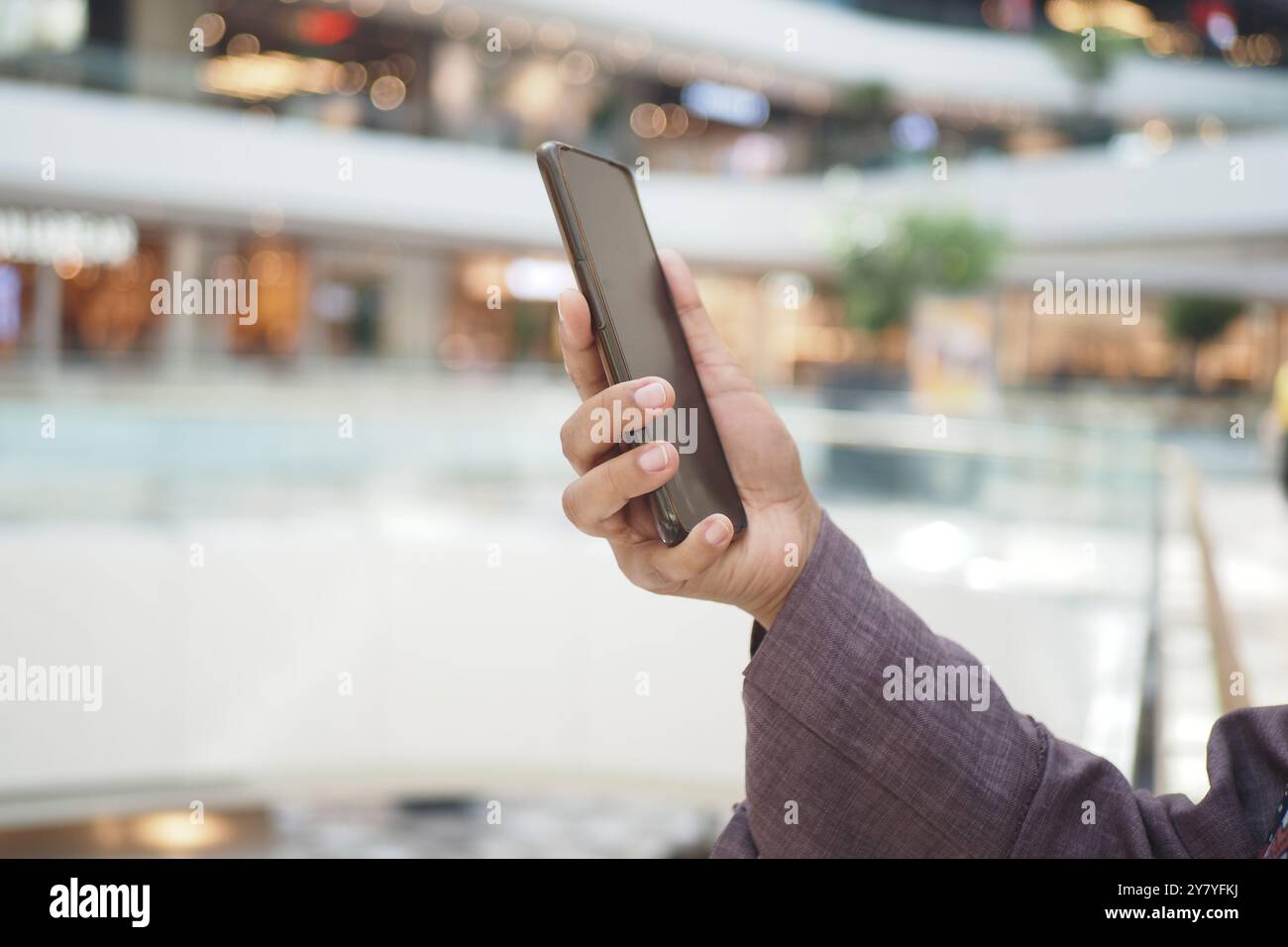 Eine Person hält ein Smartphone in der Hand, während sie sich in einem modernen Einkaufszentrum aufhält und an verschiedenen Aktivitäten teilnimmt Stockfoto