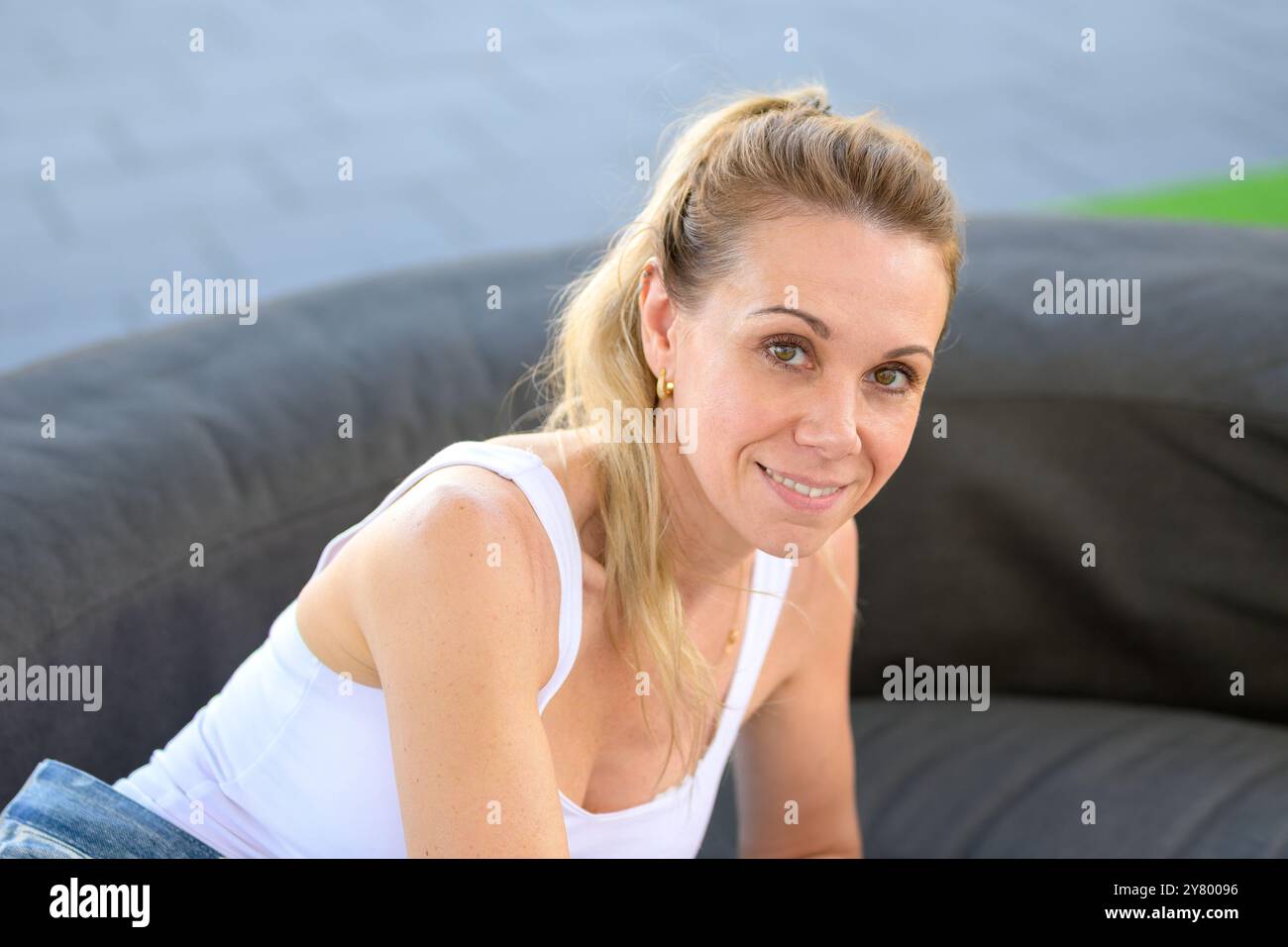 Eine Frau mit einem warmen Lächeln sitzt bequem im Freien, ihr Haar wird in einem Pferdeschwanz zurückgezogen. Stockfoto