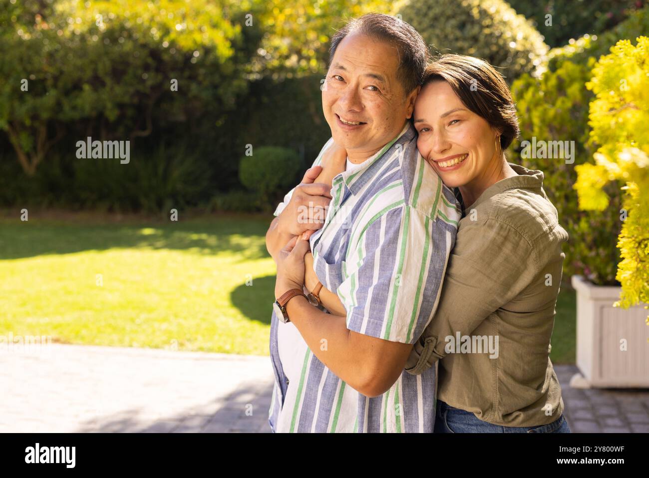 Ein Paar, lächelnde Frau mittleren Alters, umarmt asiatischen Mann draußen im sonnigen Garten und genießt Familienzeit Stockfoto