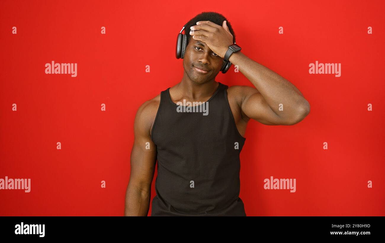 Junger afroamerikanischer Mann mit Kopfhörern steht vor einem leuchtend roten Hintergrund, der Coolness und Selbstvertrauen ausstrahlt. Stockfoto