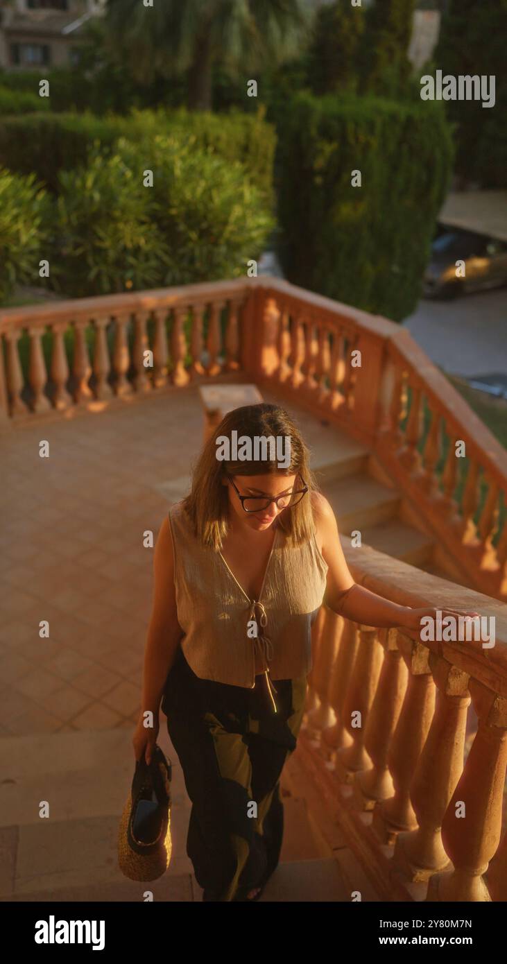 Junge hispanische Frau, die eine Außentreppe in deia, mallorca, spanien, unter Sonnenuntergangsbeleuchtung mit üppigem Garten und Palmen im Hintergrund hinaufgeht Stockfoto