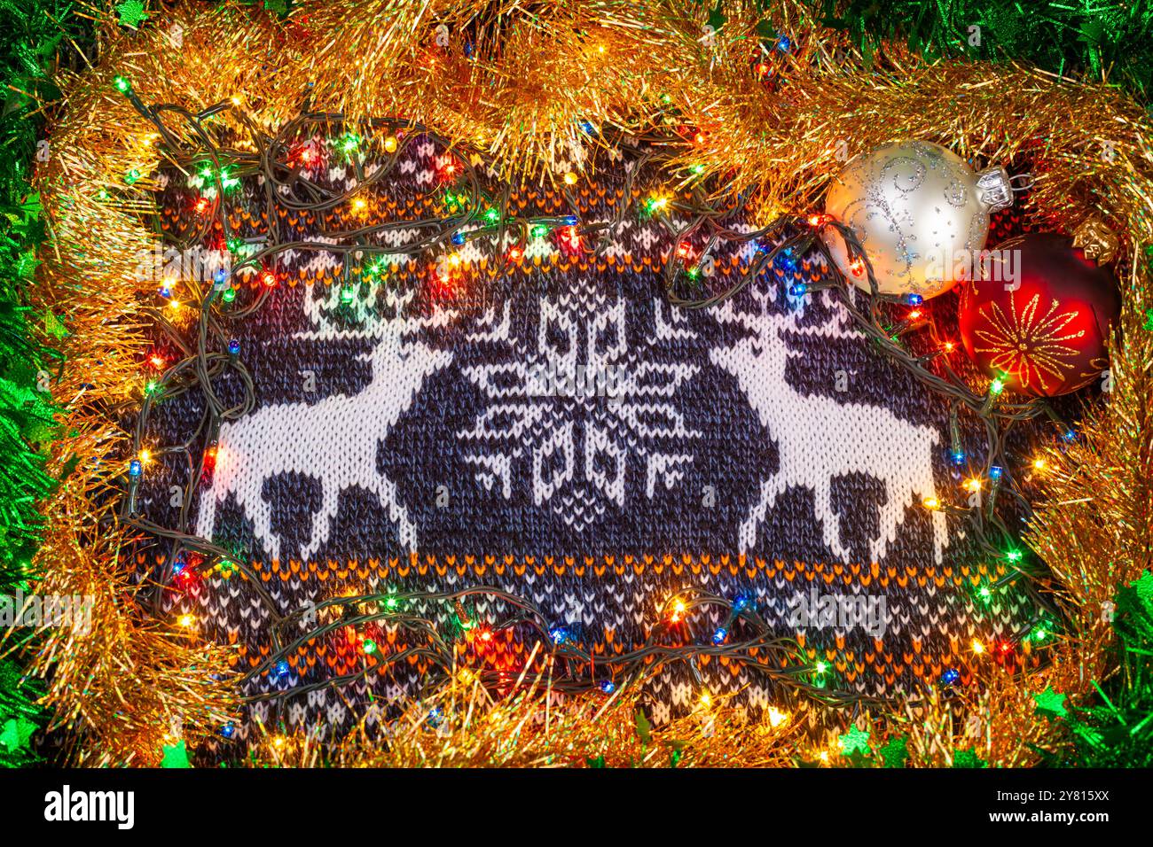 Strickmaterial mit Rentier- und Schneeflockenmuster, umgeben von farbenfrohen Lichtern, Lametta und Weihnachtsschmuck für ein festliches Arrangement Stockfoto
